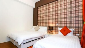 Habitación de hotel con dos camas, un ventanal grande con cortina escocesa, una mesita de noche con lámpara y un cojín rojo decorativo.