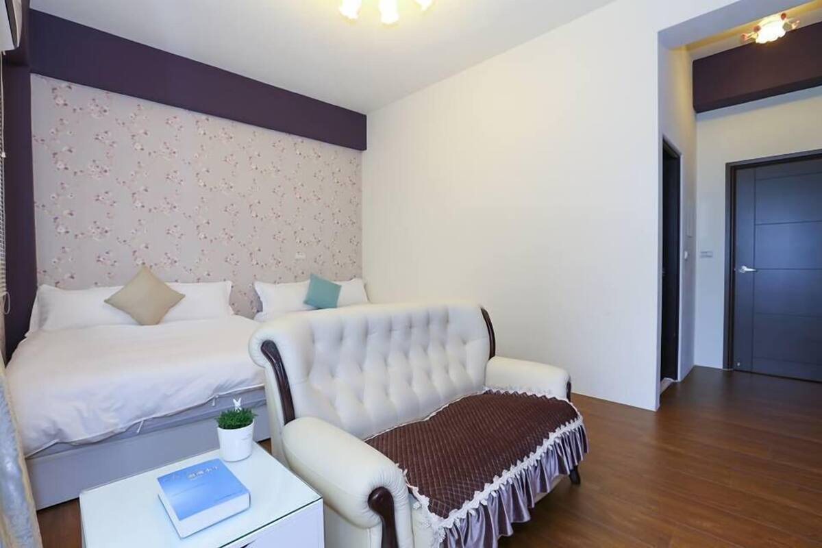 A bedroom with a bed, a sofa, a small table with a book, and a door.