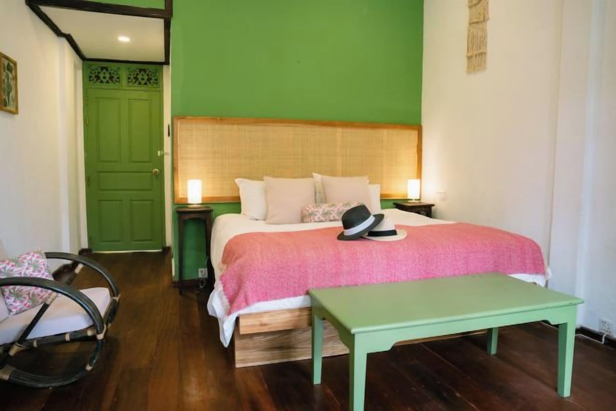 A bedroom with a green wall, a bed with a pink blanket, a wooden headboard, a nightstand with a lamp, and a green bench.