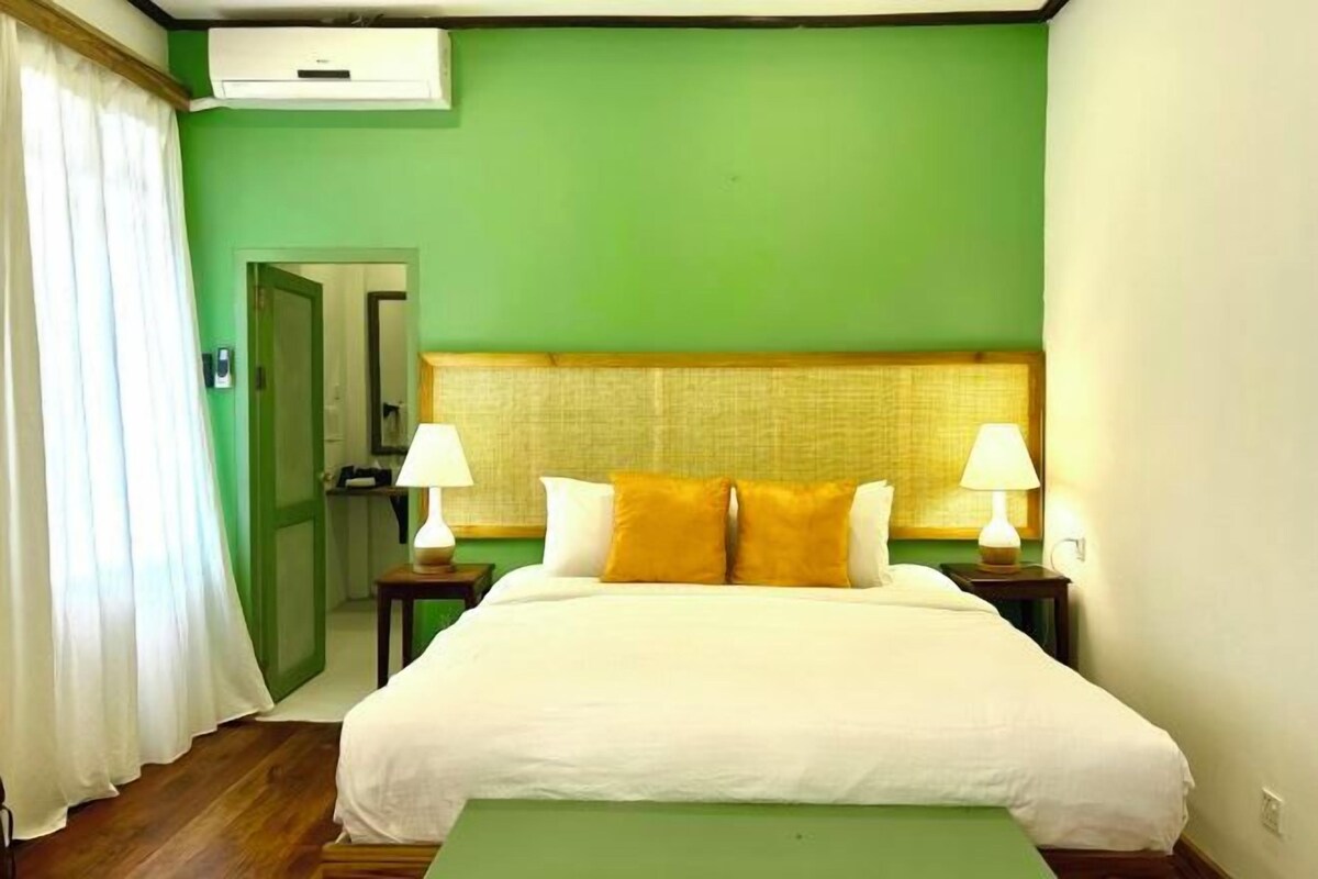 A hotel room with a green wall, a large bed with white bedding and two yellow pillows, two bedside tables with lamps, and a wooden floor.