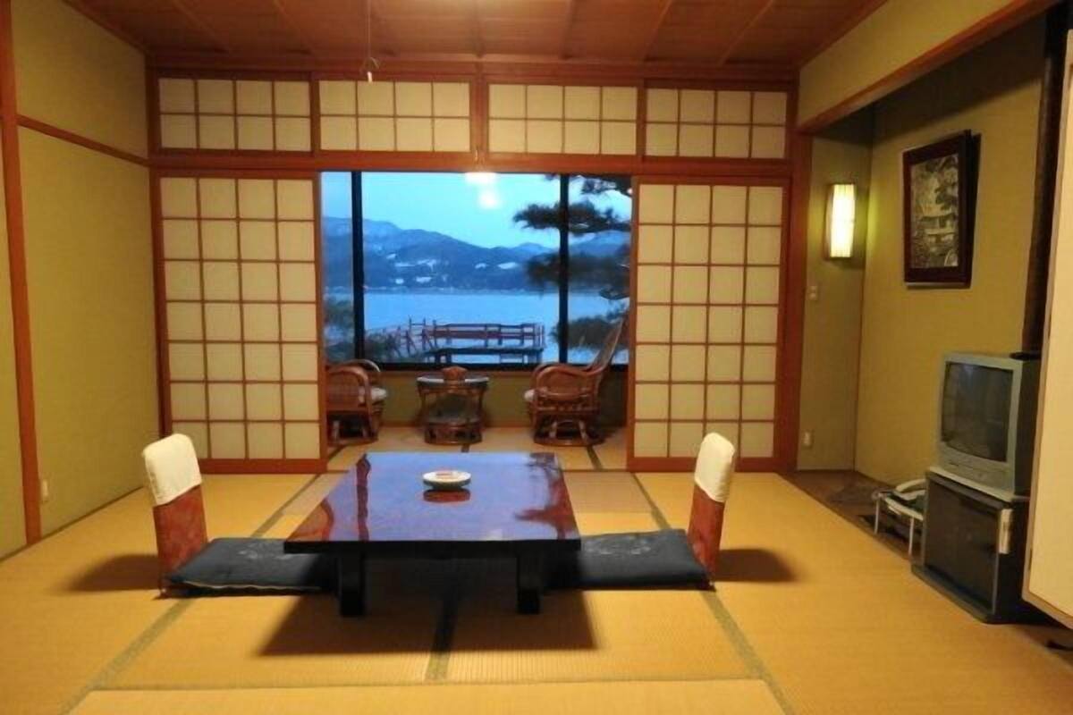 A traditional Japanese room with tatami flooring, a low table with cushions, and a view of a mountainous landscape through a large window.