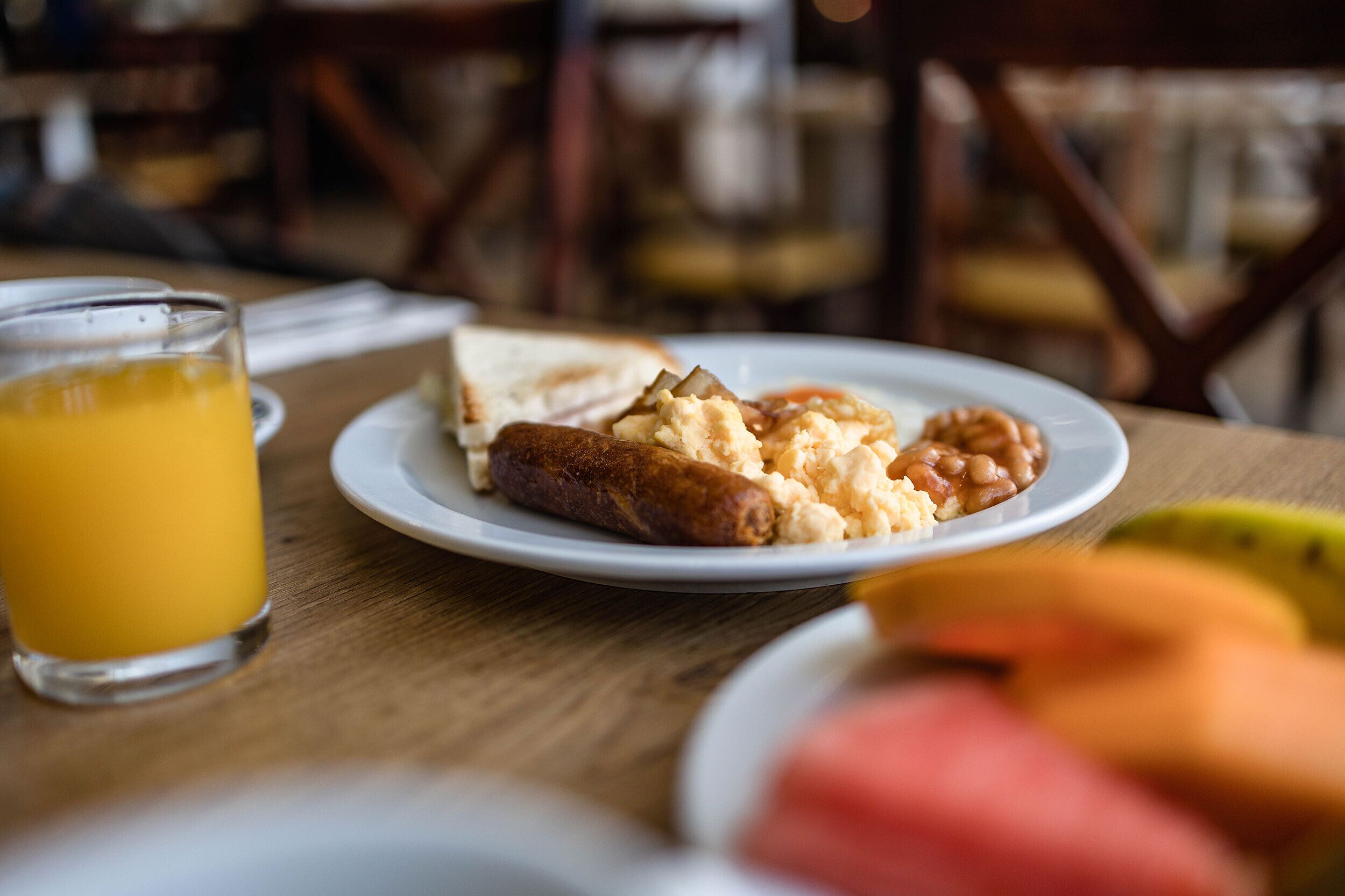 A breakfast plate with sausage, scrambled eggs, and beans, a glass of orange juice, and a banana on a wooden table.