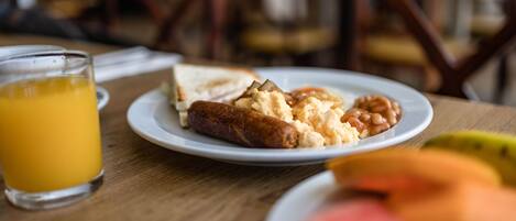 A breakfast plate with sausage, scrambled eggs, and beans, a glass of orange juice, and a banana on a wooden table.