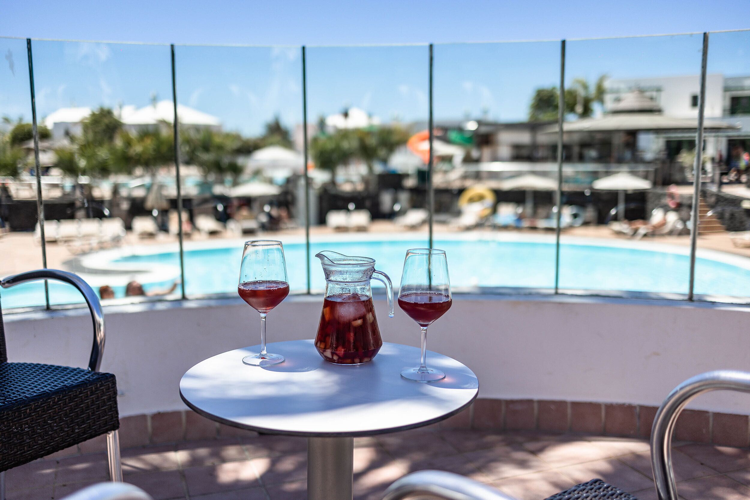 A balcony with a table, two glasses of red drink, and a pitcher, overlooking a pool area with lounge chairs and umbrellas.