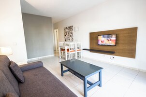 Una sala de estar moderna con un sofá, una mesa de centro, un televisor montado en la pared y una zona de comedor con sillas y una mesa.
