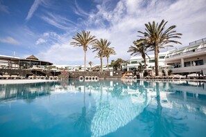 A swimming pool with lounge chairs and palm trees, surrounded by buildings with balconies. - Caybeach Sun (Yaiza)