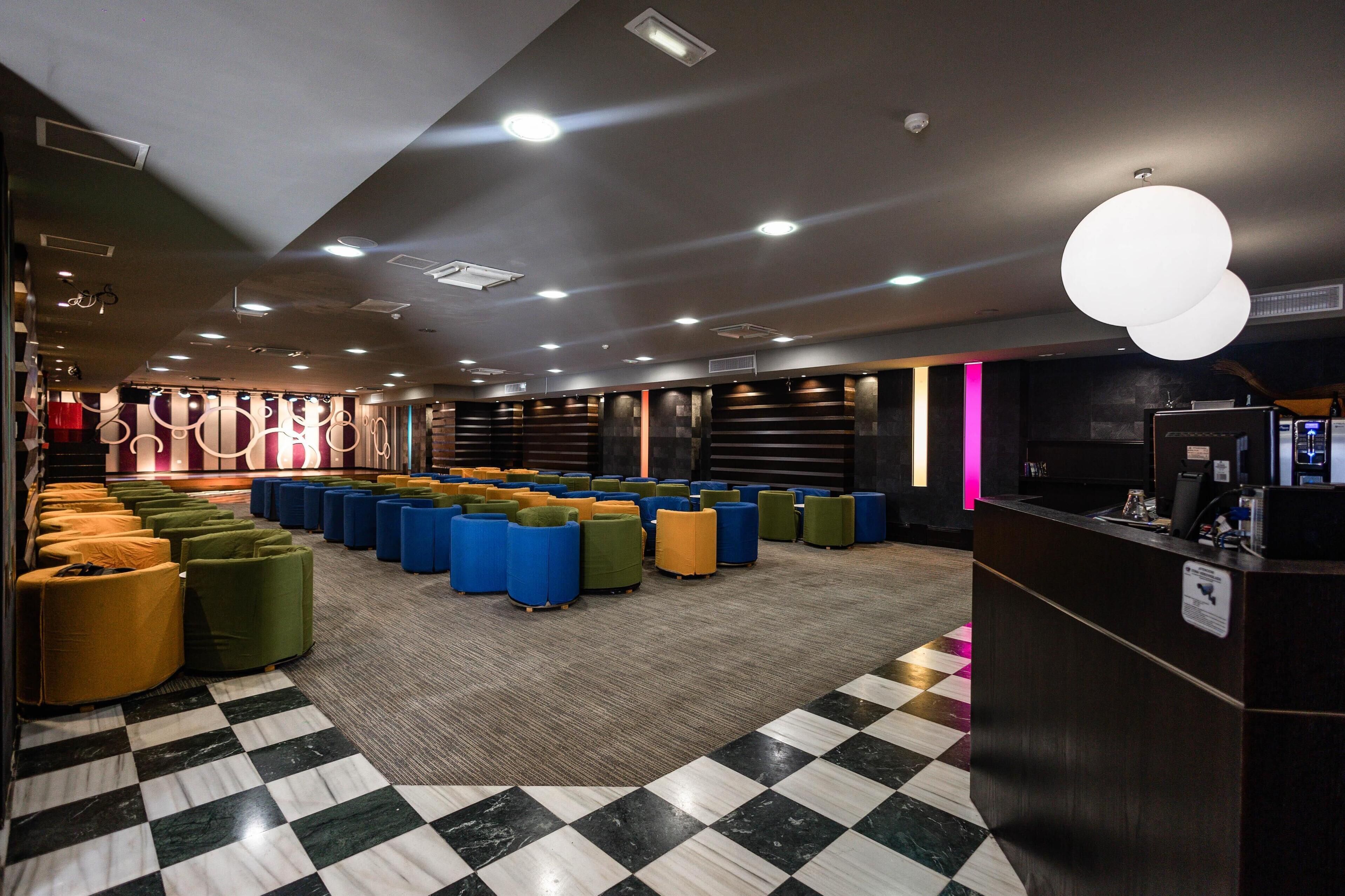 Un vestíbulo de hotel con piso a cuadros, varias zonas de estar con cojines de colores, un mostrador de recepción y un diseño de techo moderno.