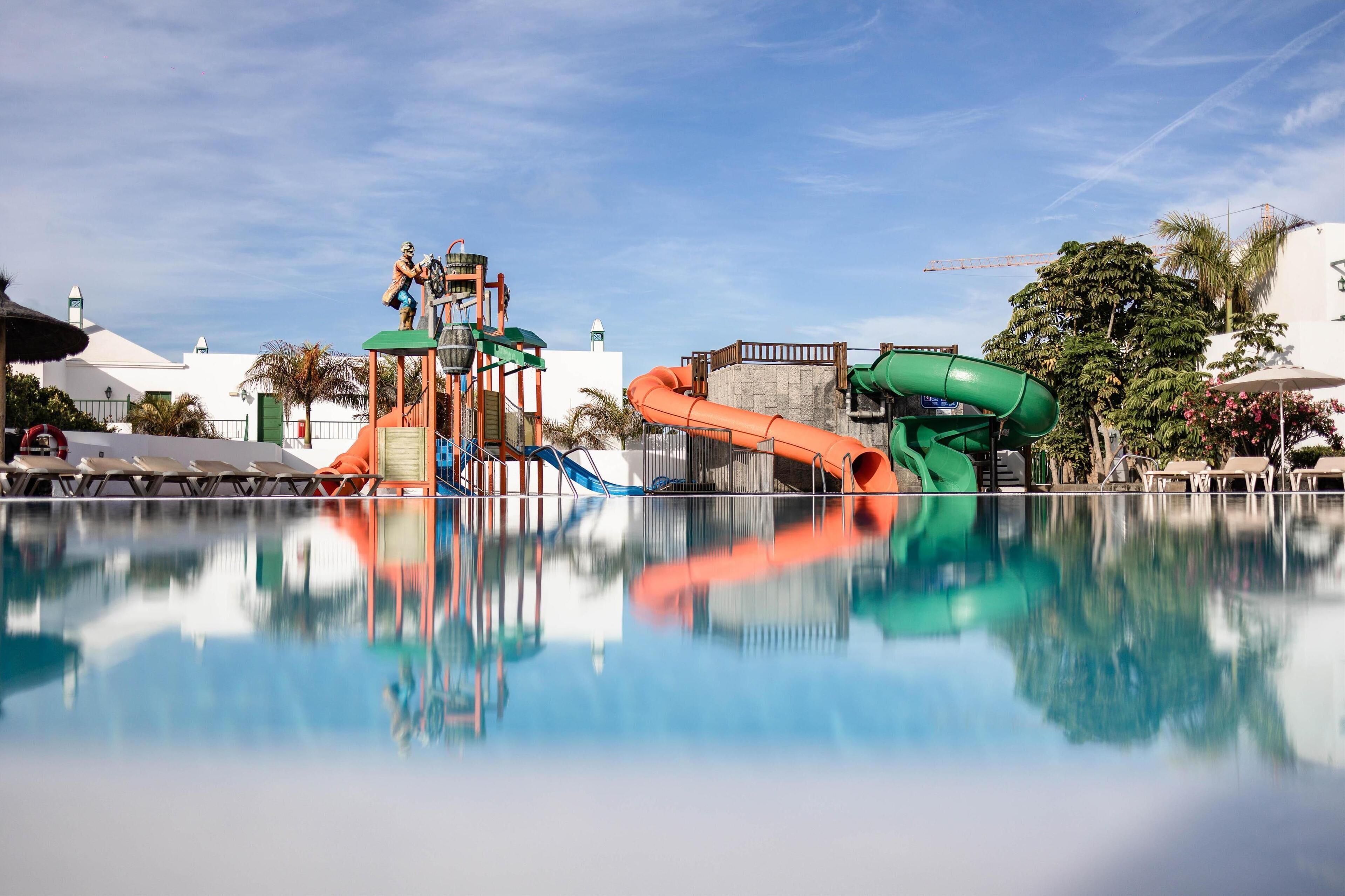 Ein Wasserpark mit Rutschen und einem Pool, der den Himmel und die Gebäude spiegelt.