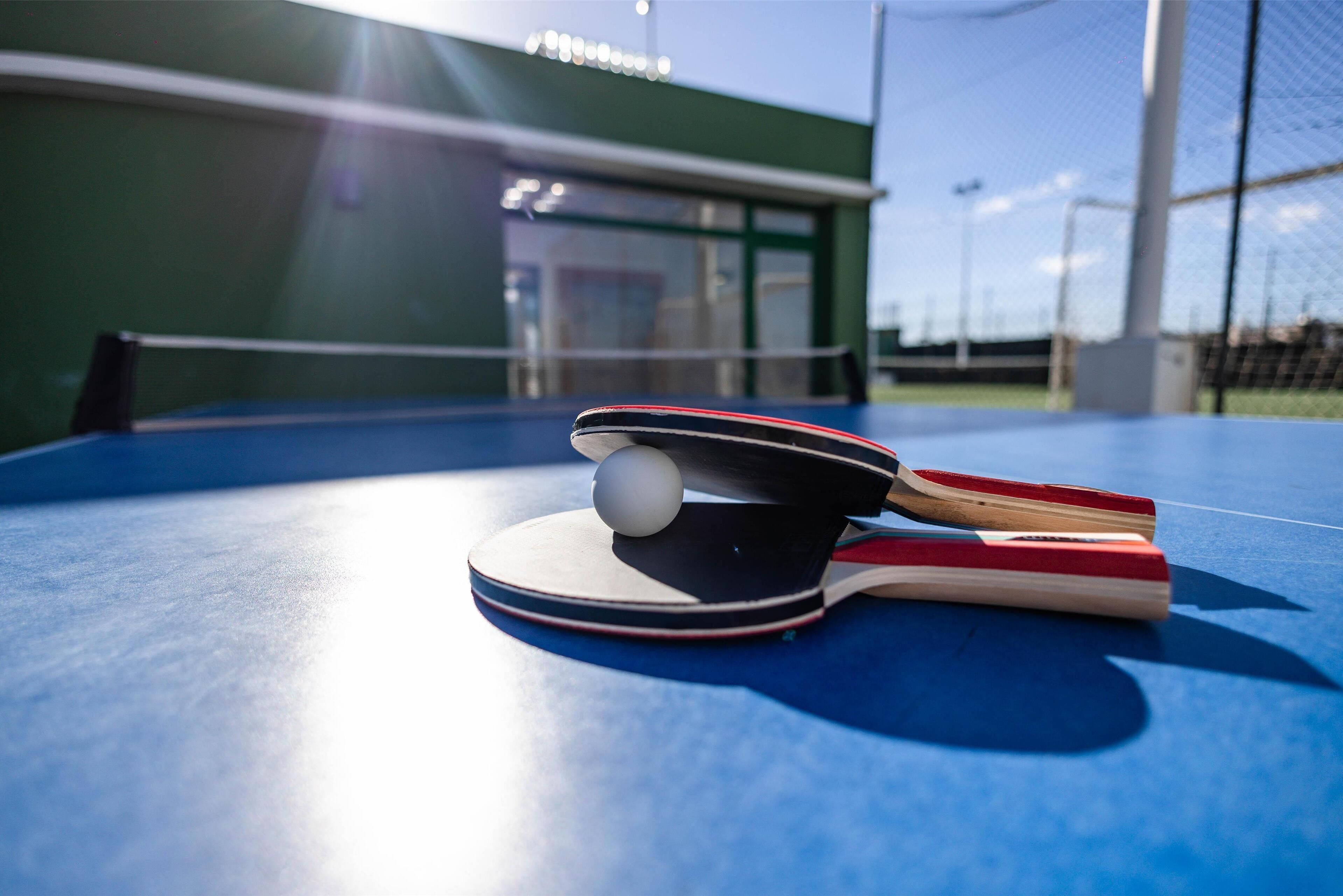 A ping pong table with paddles and a ball, outdoors with a building in the background.