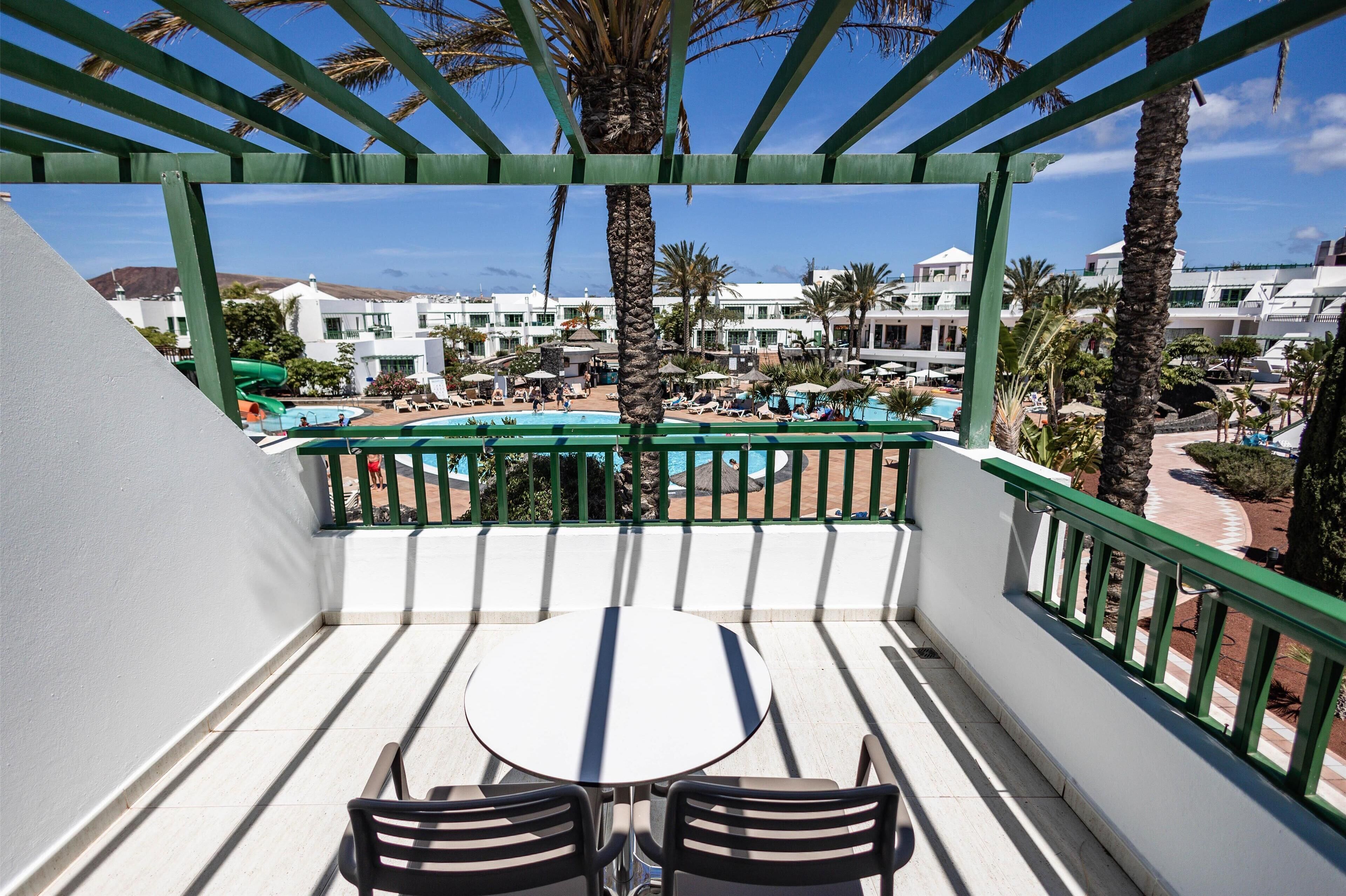 Un balcón con mesa y sillas, con vistas a una piscina rodeada de palmeras y edificios del complejo.