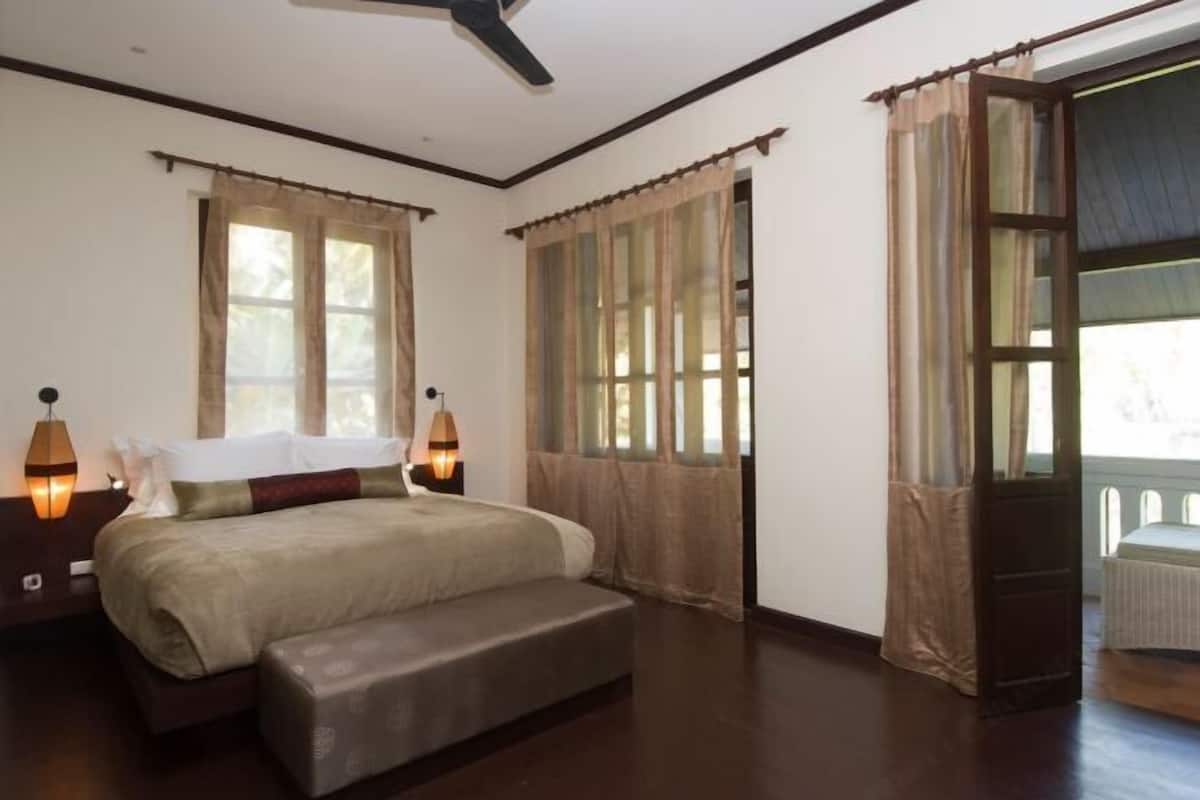 A bedroom with a bed, bedside lamps, a bench, a ceiling fan, and large windows with curtains.