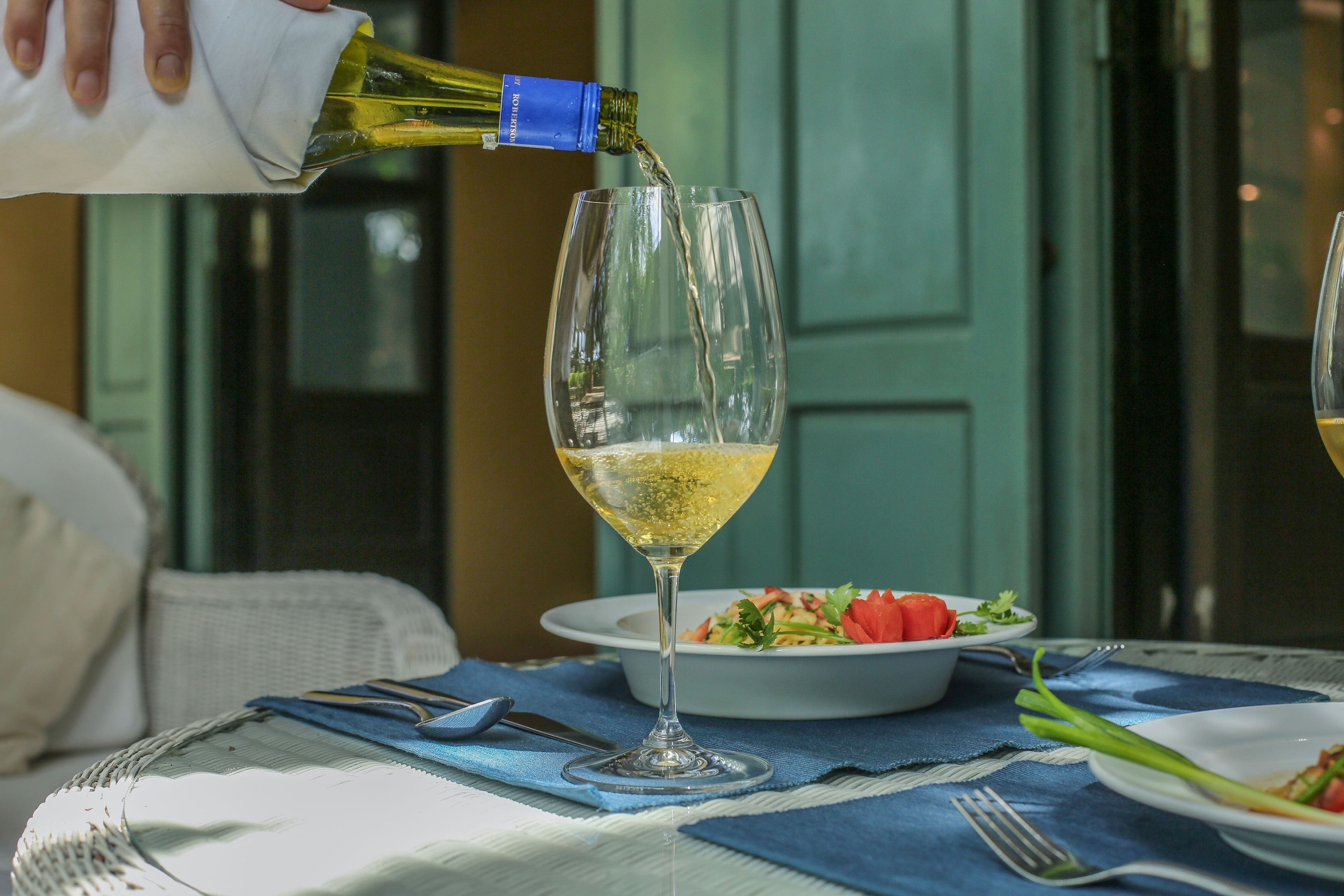 A table with a wine glass being filled, a bowl of salad, and a wicker chair.