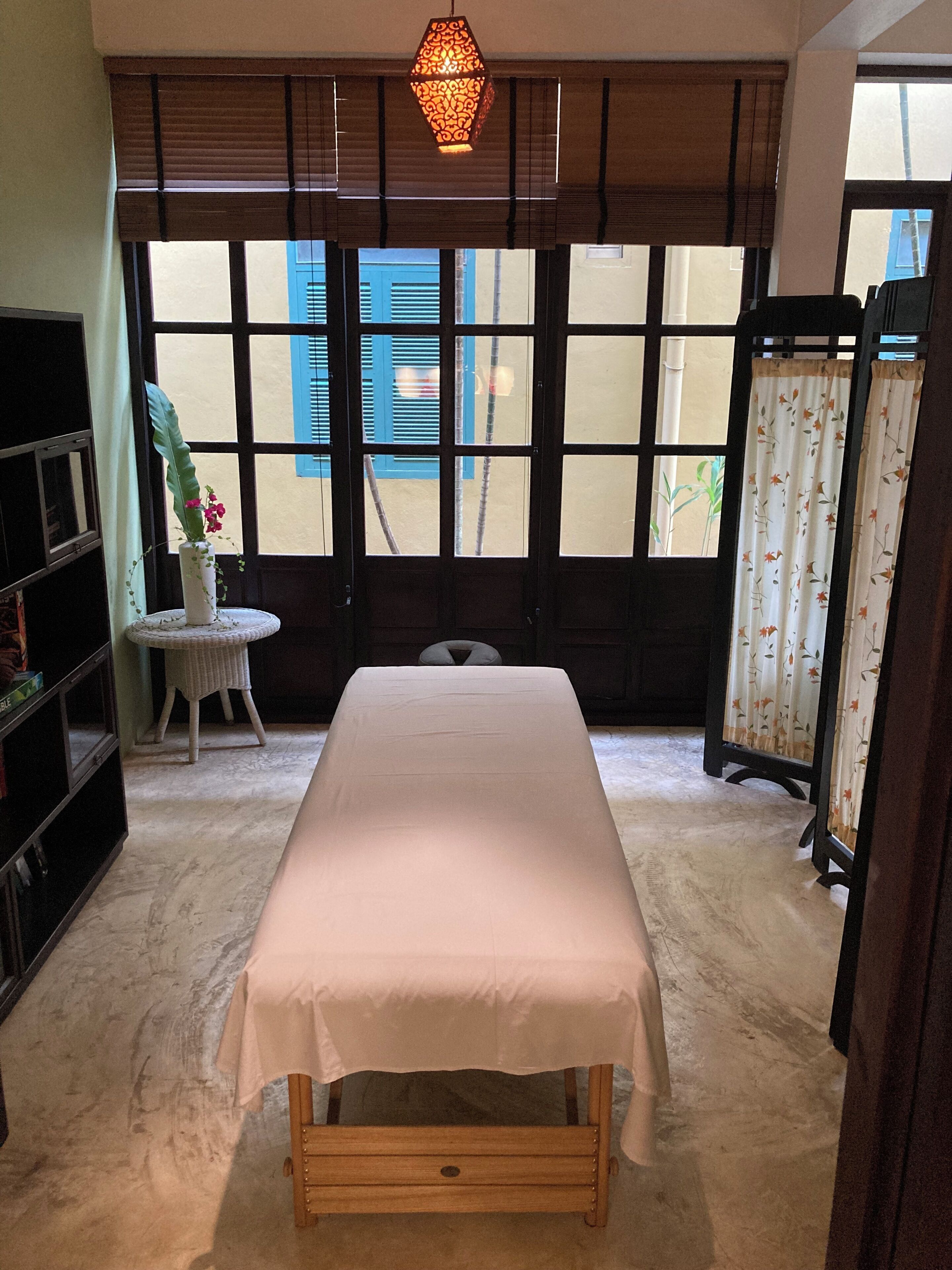 A room with a bed, a small round table with a chair, a bookshelf, and a window with blinds.