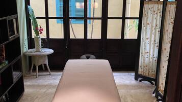 A room with a bed, a small round table with a chair, a bookshelf, and a window with blinds.