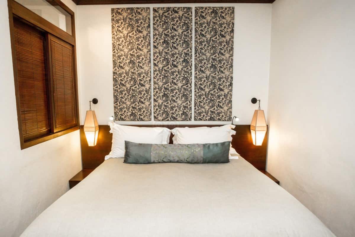 A bedroom with a bed, two bedside lamps, a window with blinds, and a patterned wall hanging.