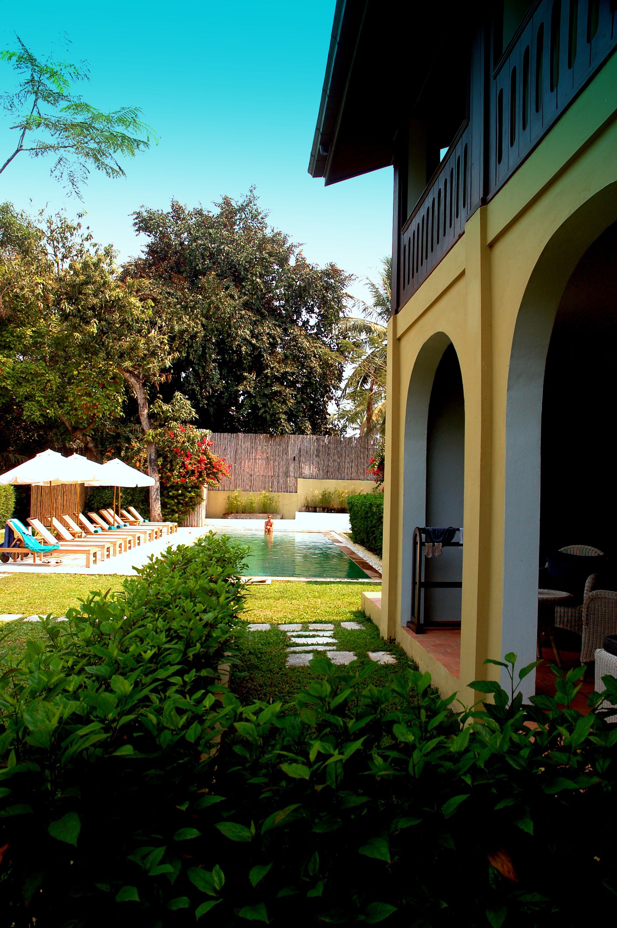 A pool area with lounge chairs and umbrellas, surrounded by greenery and a wooden fence.