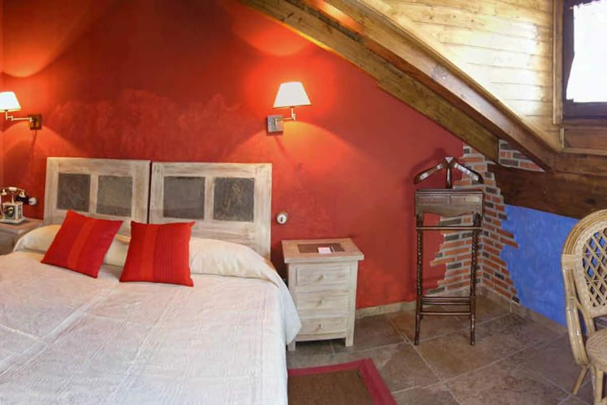 A bedroom with a wooden ceiling, a bed with red pillows, a small table with a chessboard, and a chair.