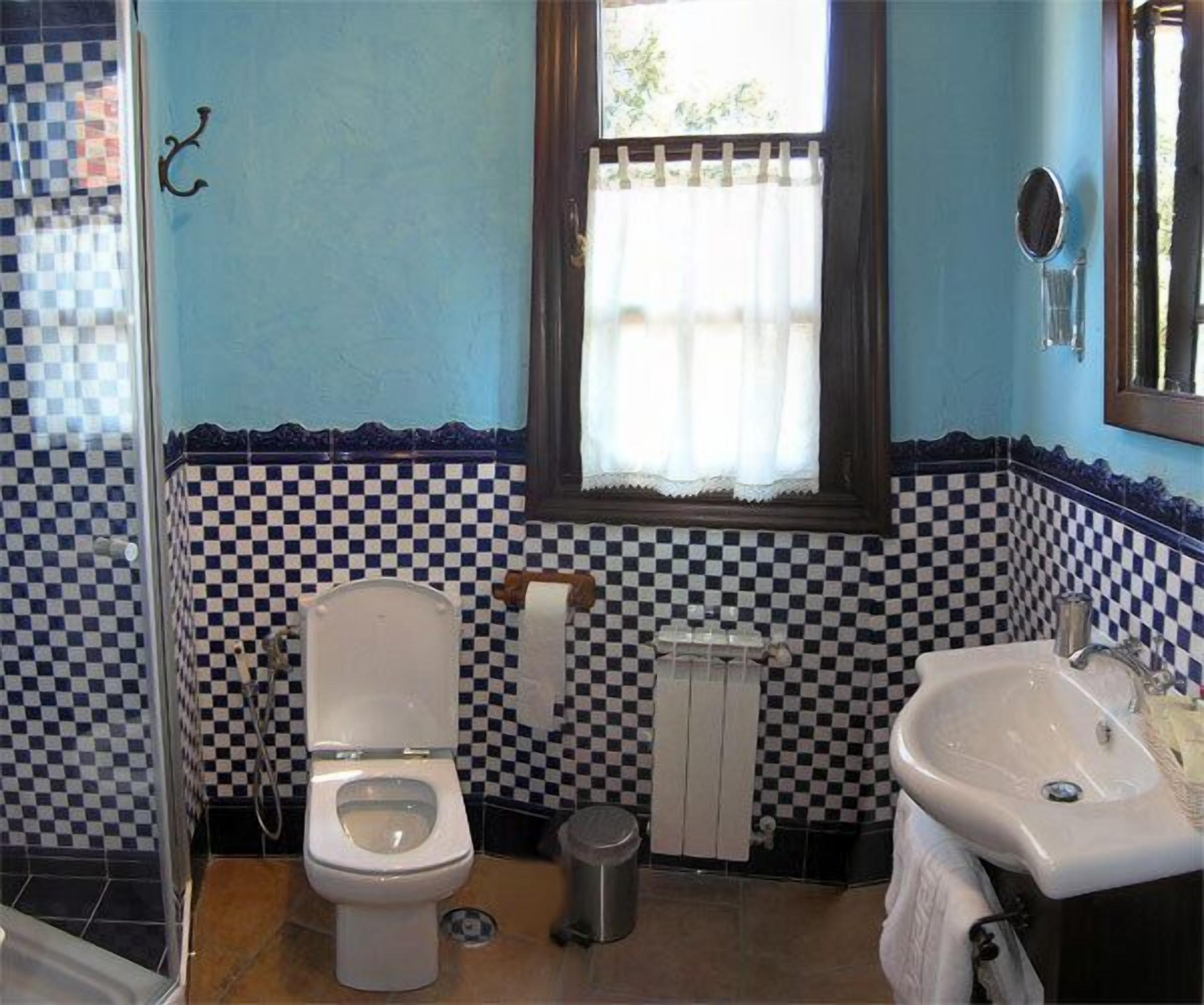 Un baño con pared de baldosas a cuadros, inodoro, lavamanos y ventana con cortina.