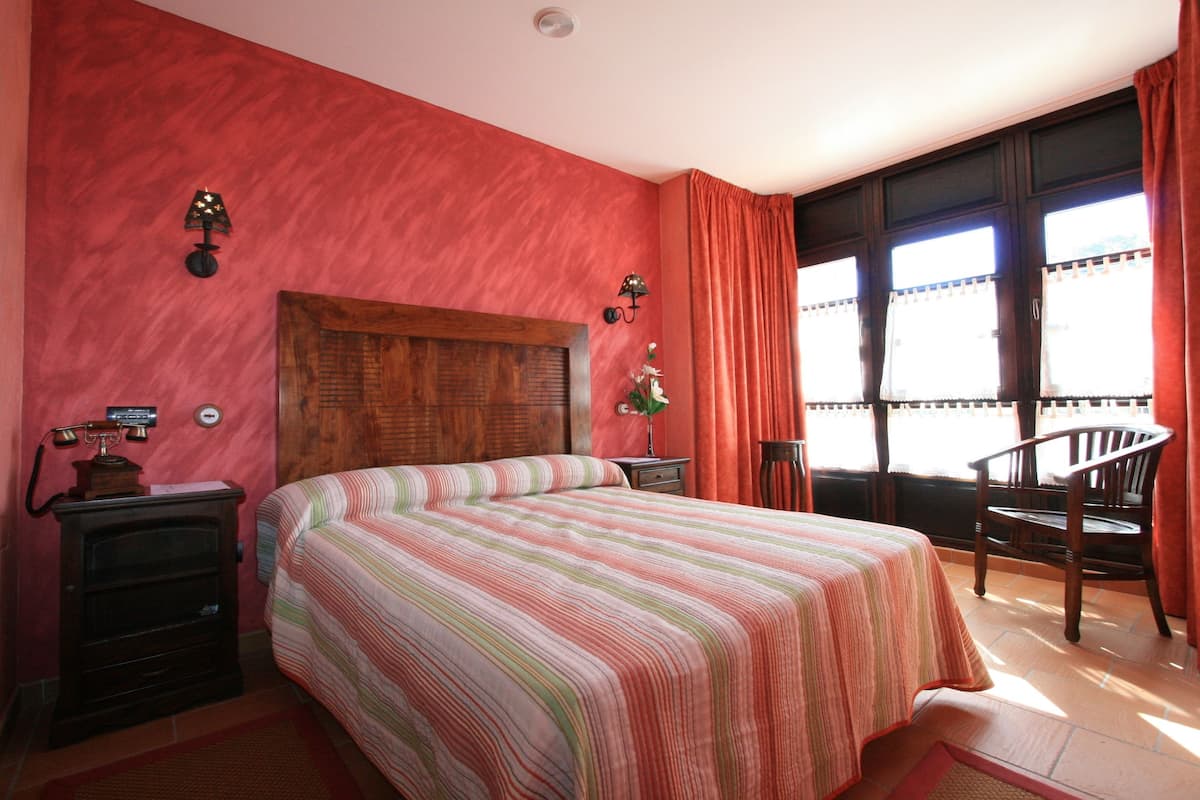 A bedroom with a large bed, wooden headboard, two bedside tables, a nightstand, a chair, and a window with curtains.