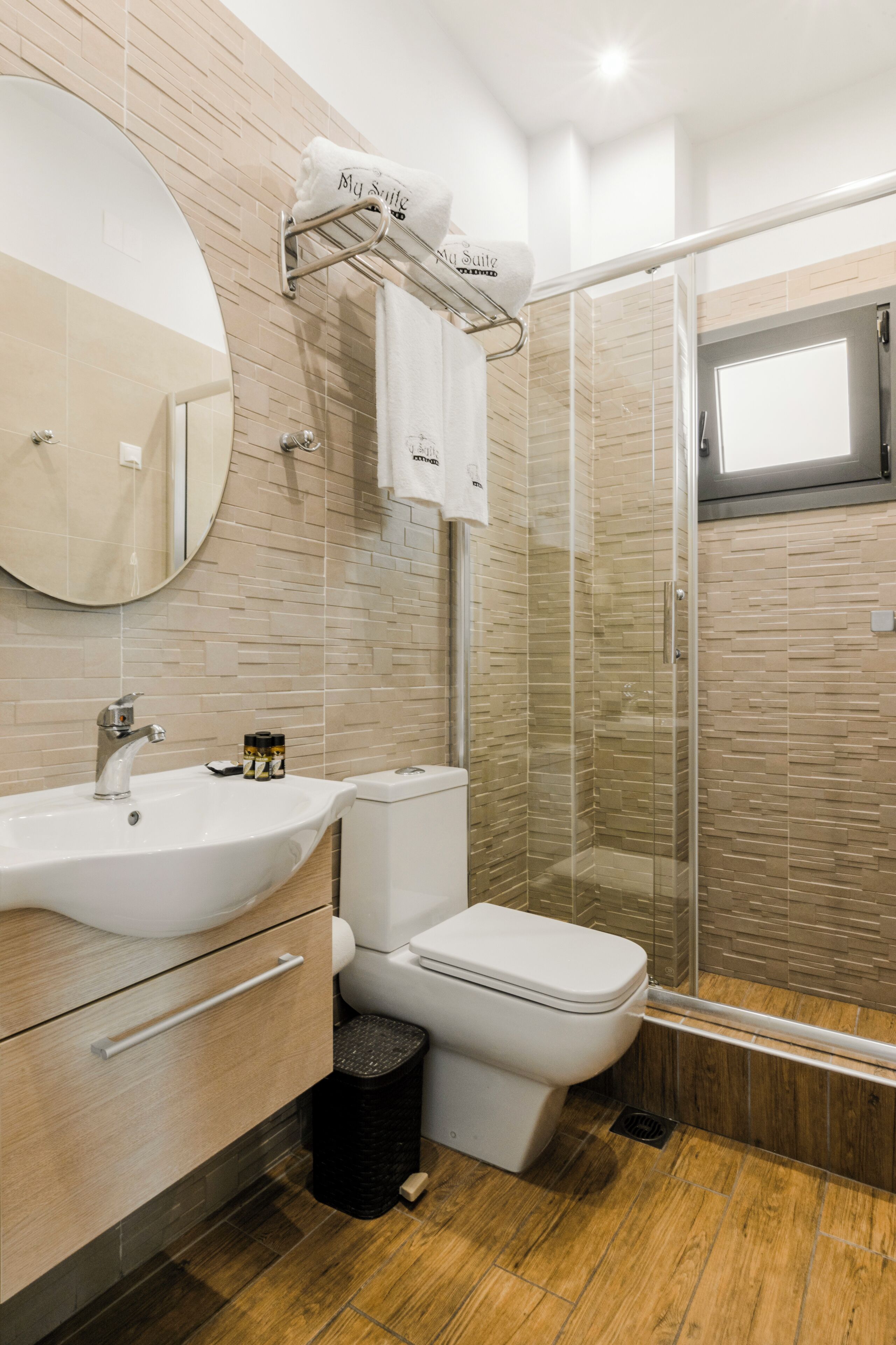 A modern bathroom with a beige tiled wall, a round mirror, a white sink, a toilet, and a glass shower enclosure.