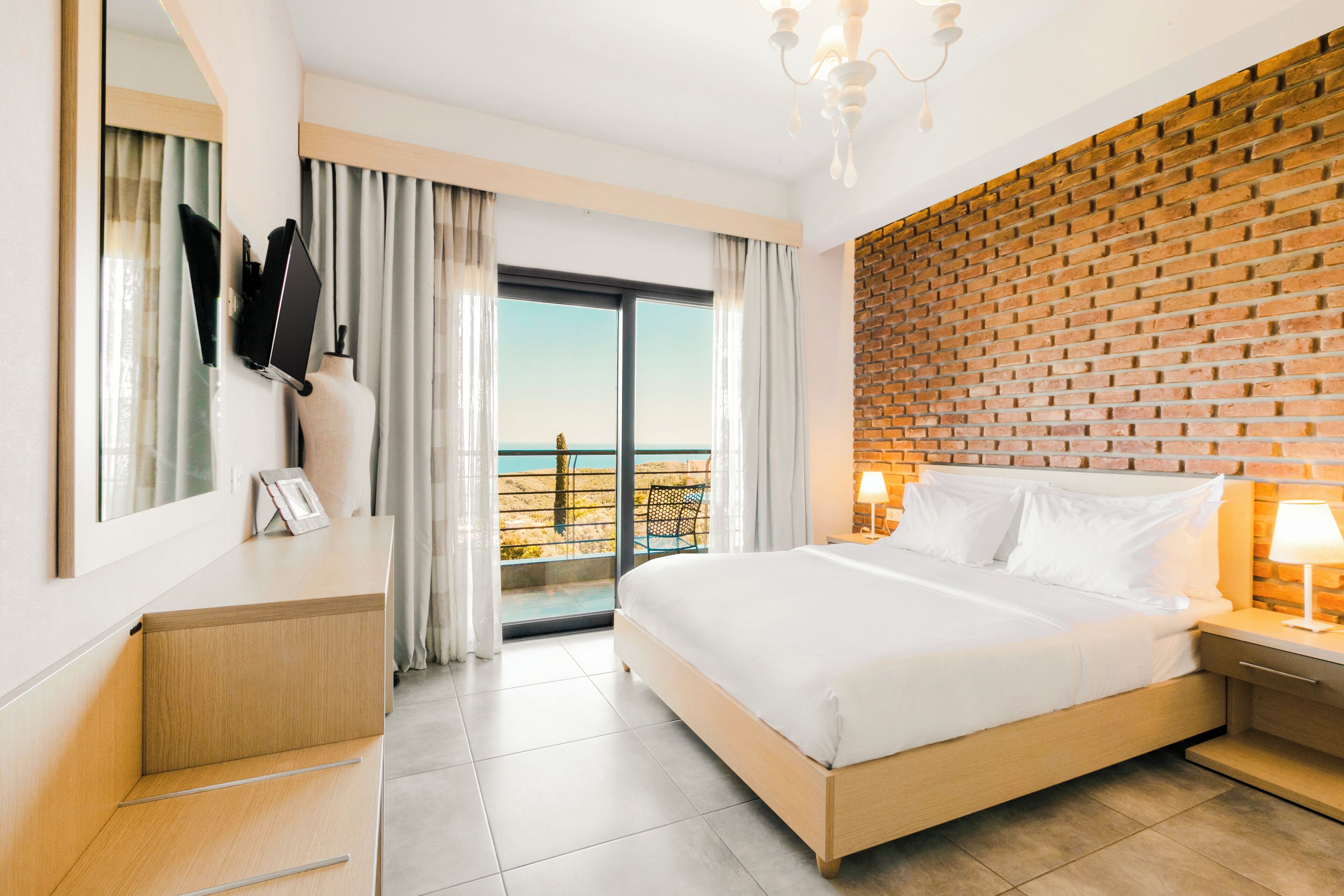 A hotel room with a large bed, a wooden desk, a mirror, a nightstand with a lamp, and a view of the sea through a sliding door.