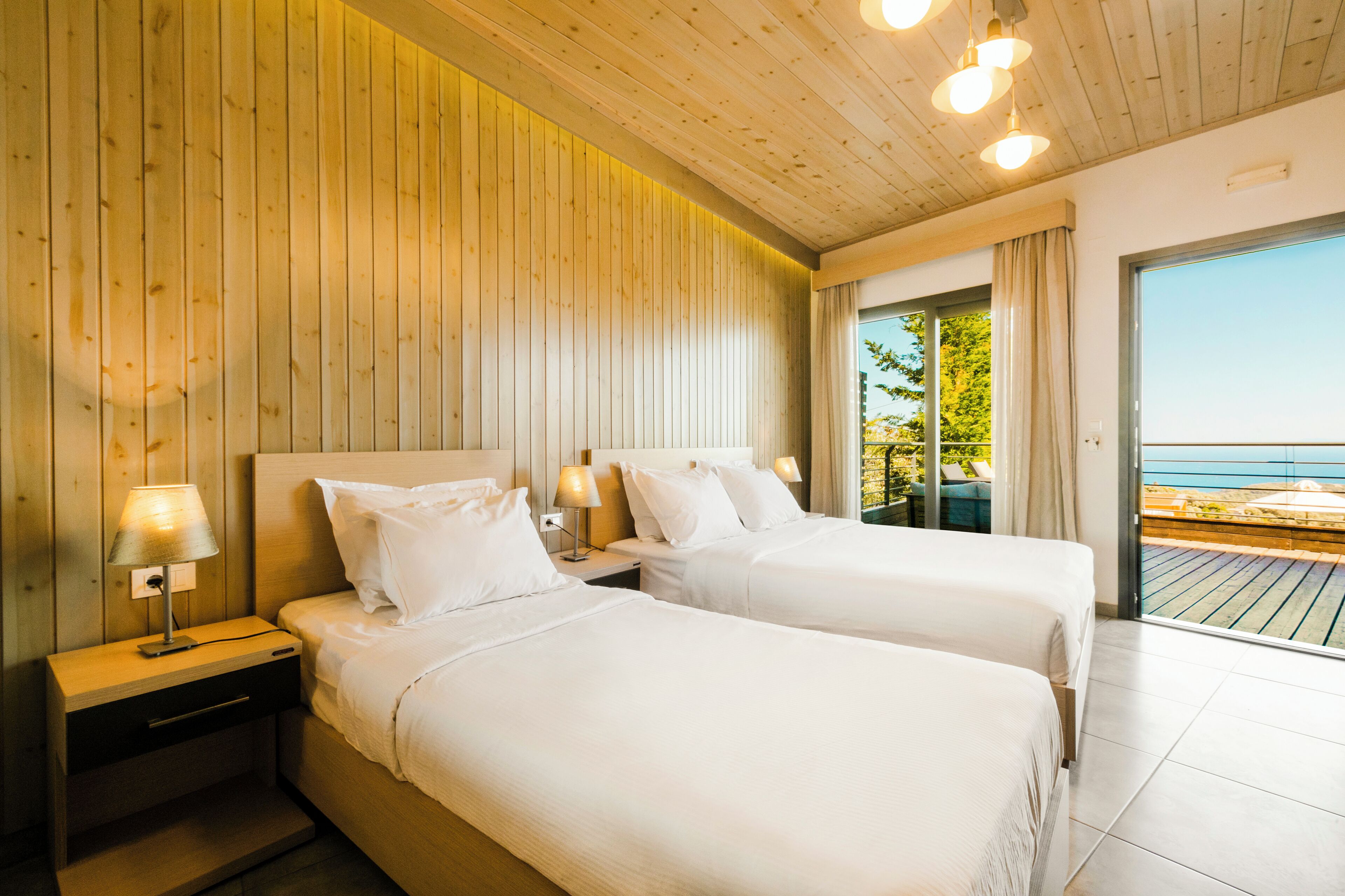 A hotel room with two beds, wooden paneling on the walls, and a balcony with a view of the sea.
