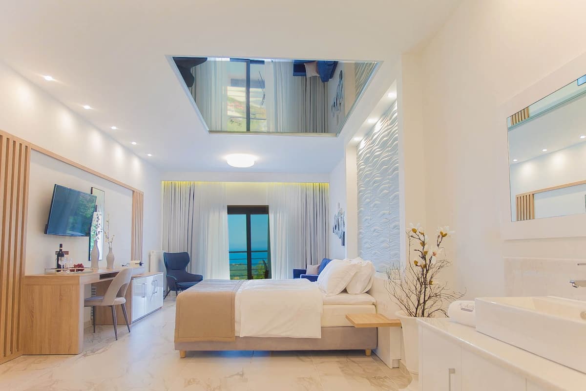 A modern hotel room with a bed, a desk, a chair, a large mirror, a bathtub, and a view of the sea.