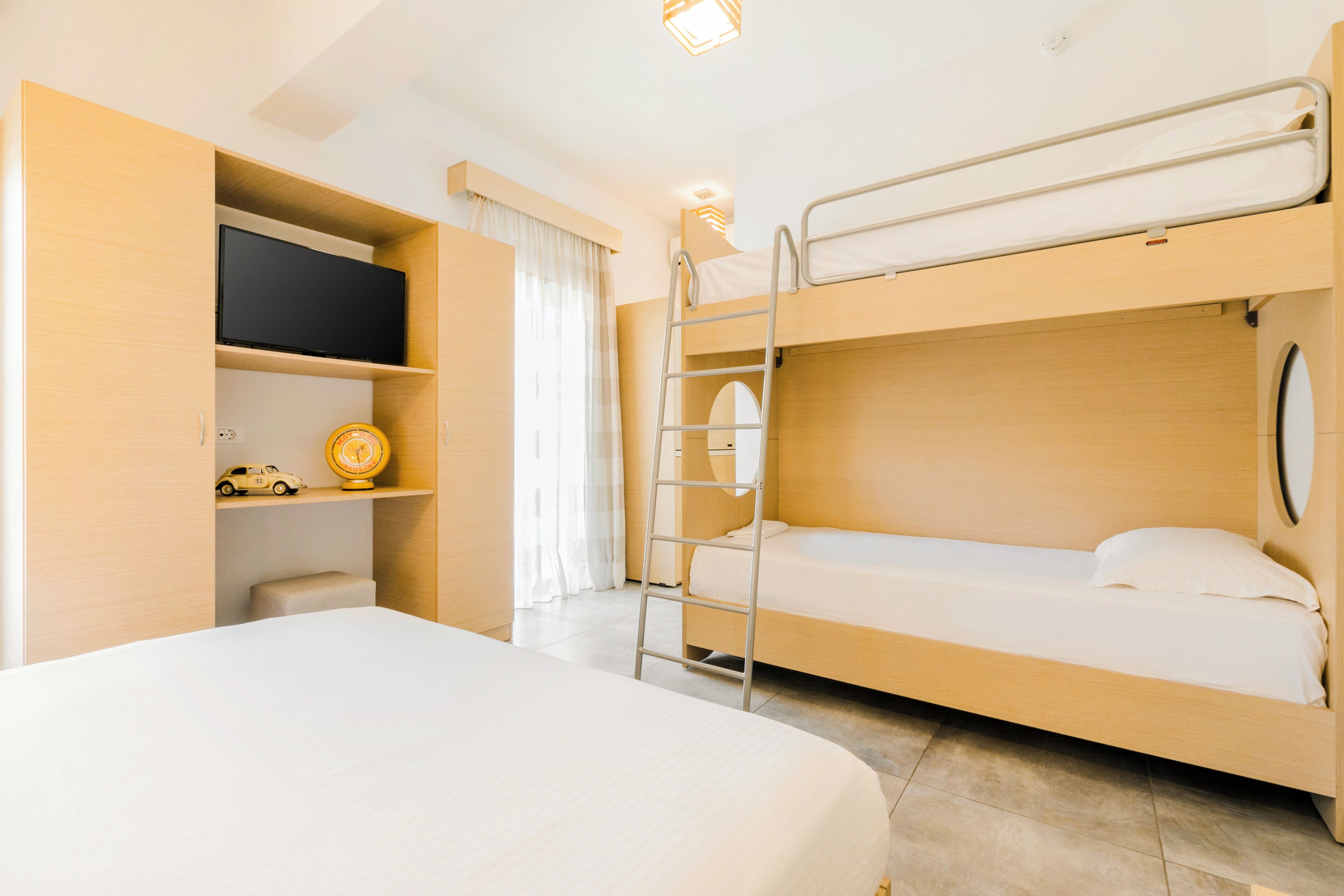 A hotel room with a bunk bed, a single bed, a TV on a shelf, and a small table with a toy car and a clock.