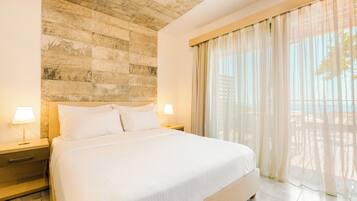 A large bed with white linens, two bedside tables with lamps, a stone wall, and a large window with sheer curtains.