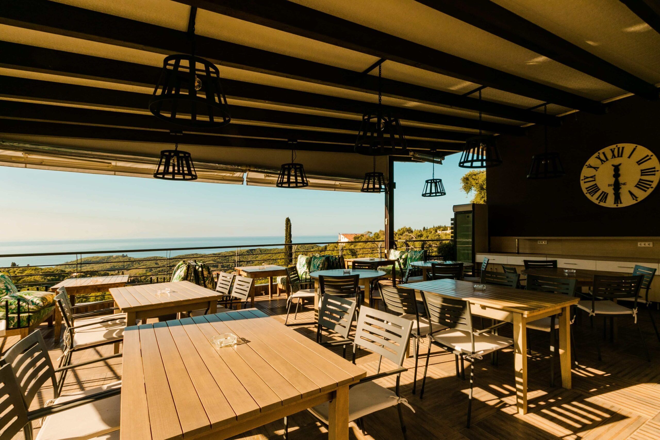 An outdoor dining area with wooden tables and chairs, a large clock on the wall, and a view of the sea.