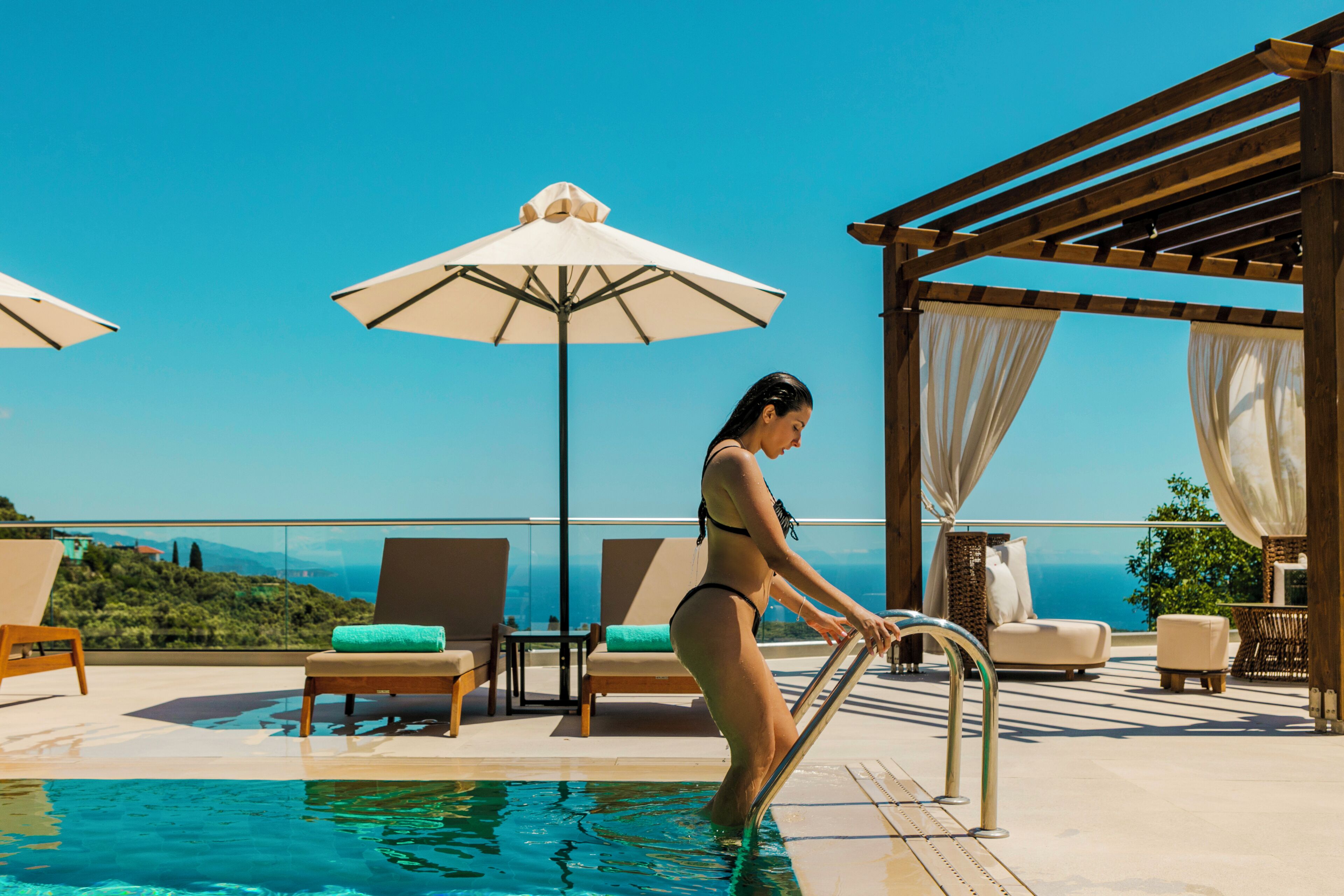 Una mujer en traje de baño sale de una piscina en una terraza en la azotea, con sillones de descanso, sombrillas y vistas al océano.