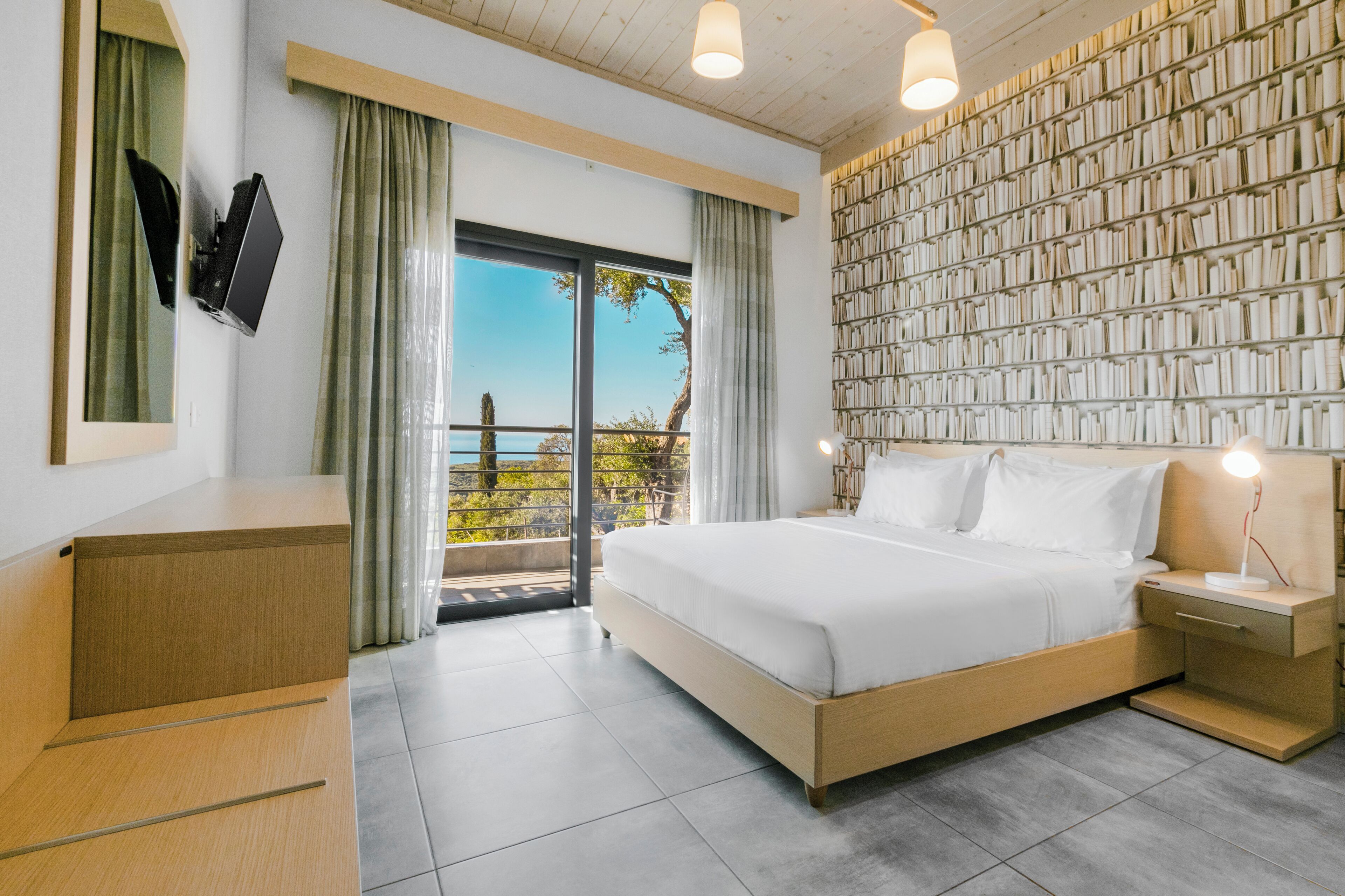 Quarto de hotel moderno com uma cama grande, televisão de tela plana na parede, mesa de cabeceira com abajur e vista para o exterior através de uma porta de vidro deslizante.