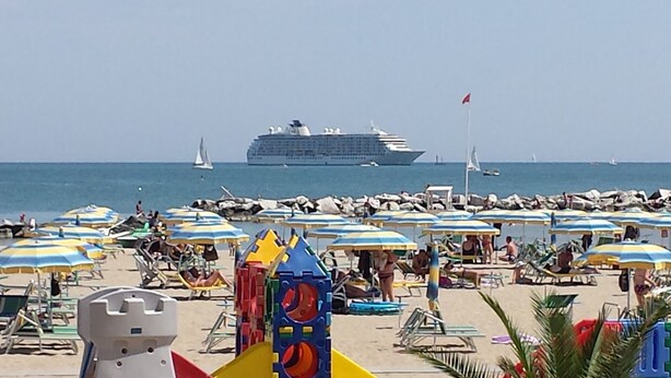 Una playa con múltiples sombrillas y sillas de descanso, un gran crucero al fondo, y una vista despejada del mar.