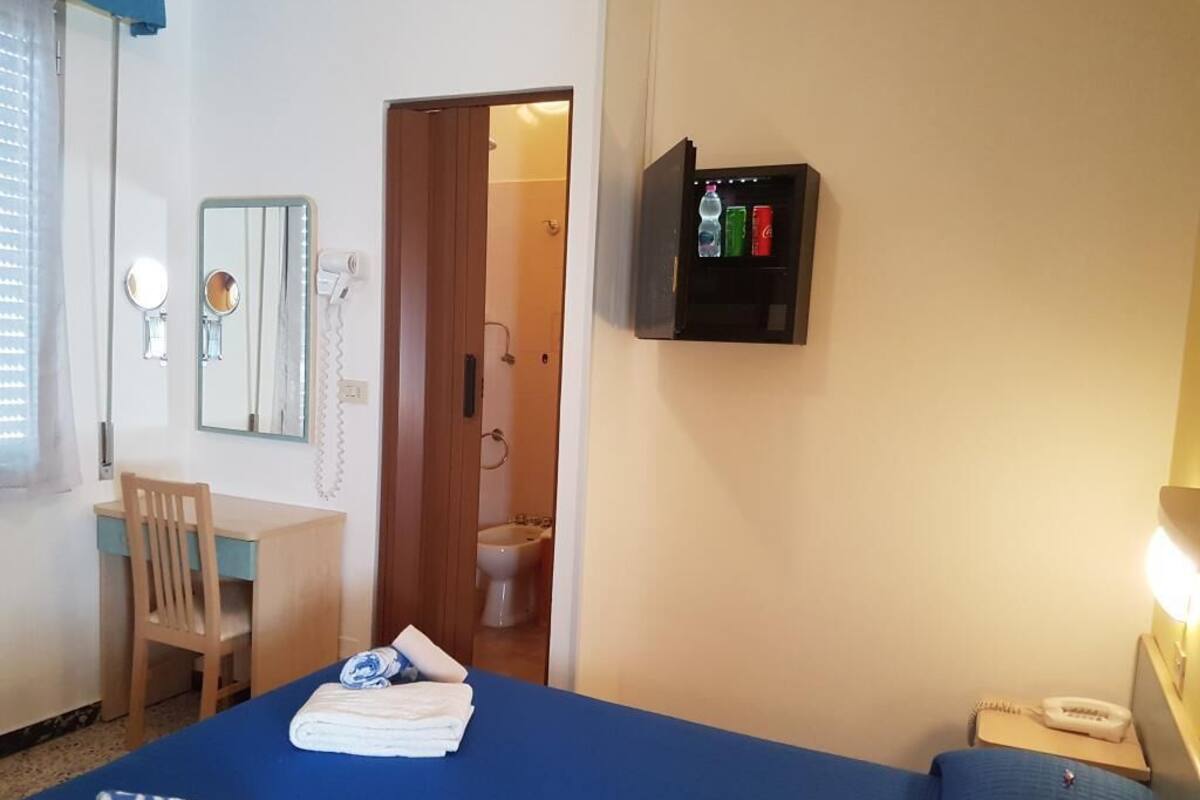 Une chambre d’hôtel avec un lit, un bureau, une chaise, un miroir, une salle de bain visible à travers une porte ouverte et un meuble à pharmacie fixé au mur.