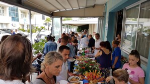 Un grupo de personas reunidas alrededor de una mesa con comida y bebidas, frente a un edificio con grandes ventanales.