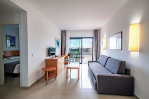 A hotel room with a bed, a sofa, a desk with a chair, a small table, and a TV. There is a window with curtains and a view of the outside. - Caybeach Papagayos (Yaiza)