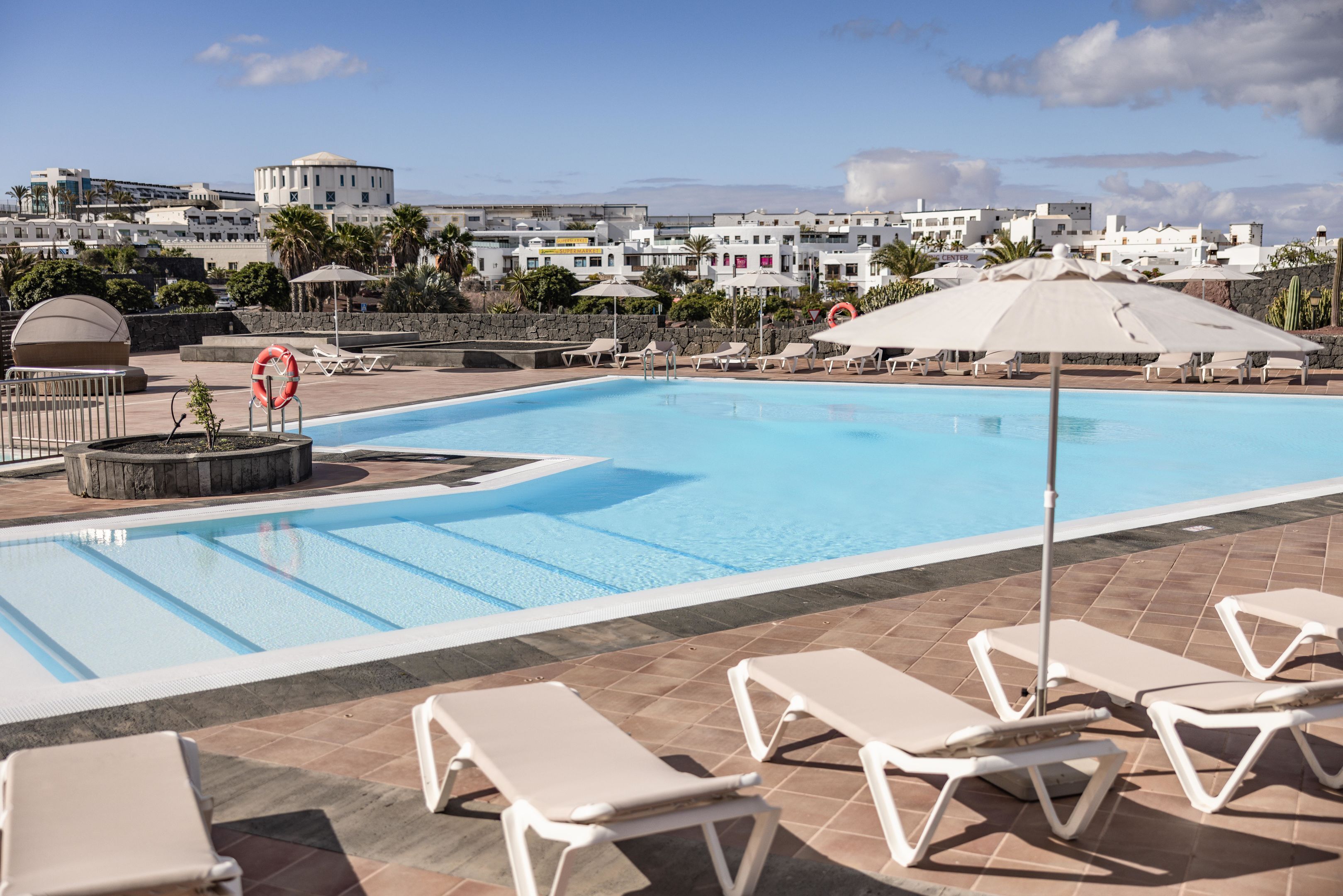 A swimming pool with lounge chairs and umbrellas, surrounded by a tiled deck and a view of buildings in the background.