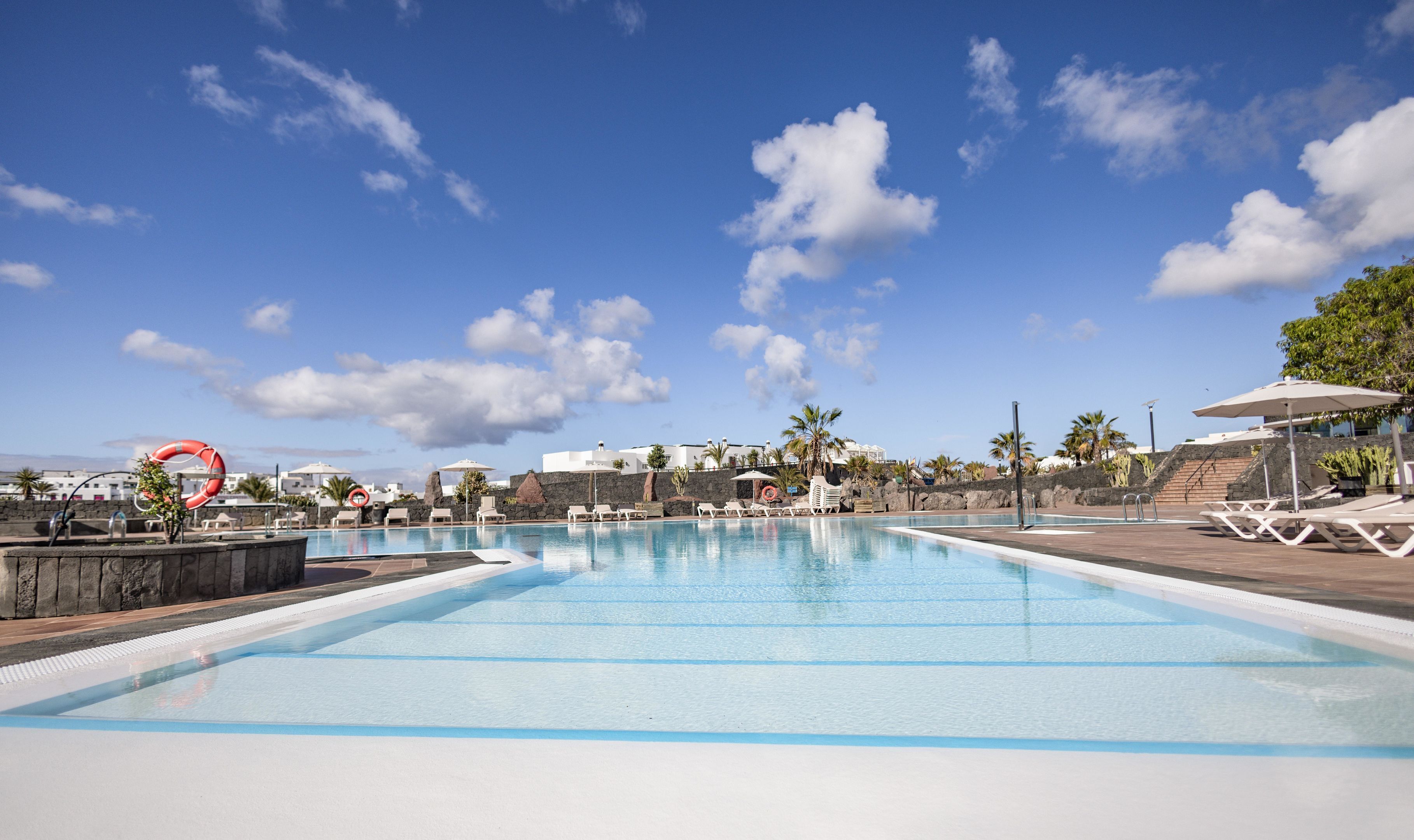 Una piscina con sillones de descanso y sombrillas, rodeada de palmeras y edificios.