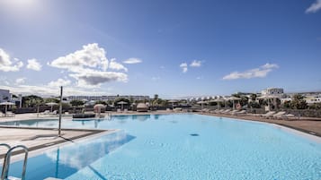 Una amplia piscina con agua cristalina, rodeada de sillones de descanso y sombrillas, con edificios al fondo.