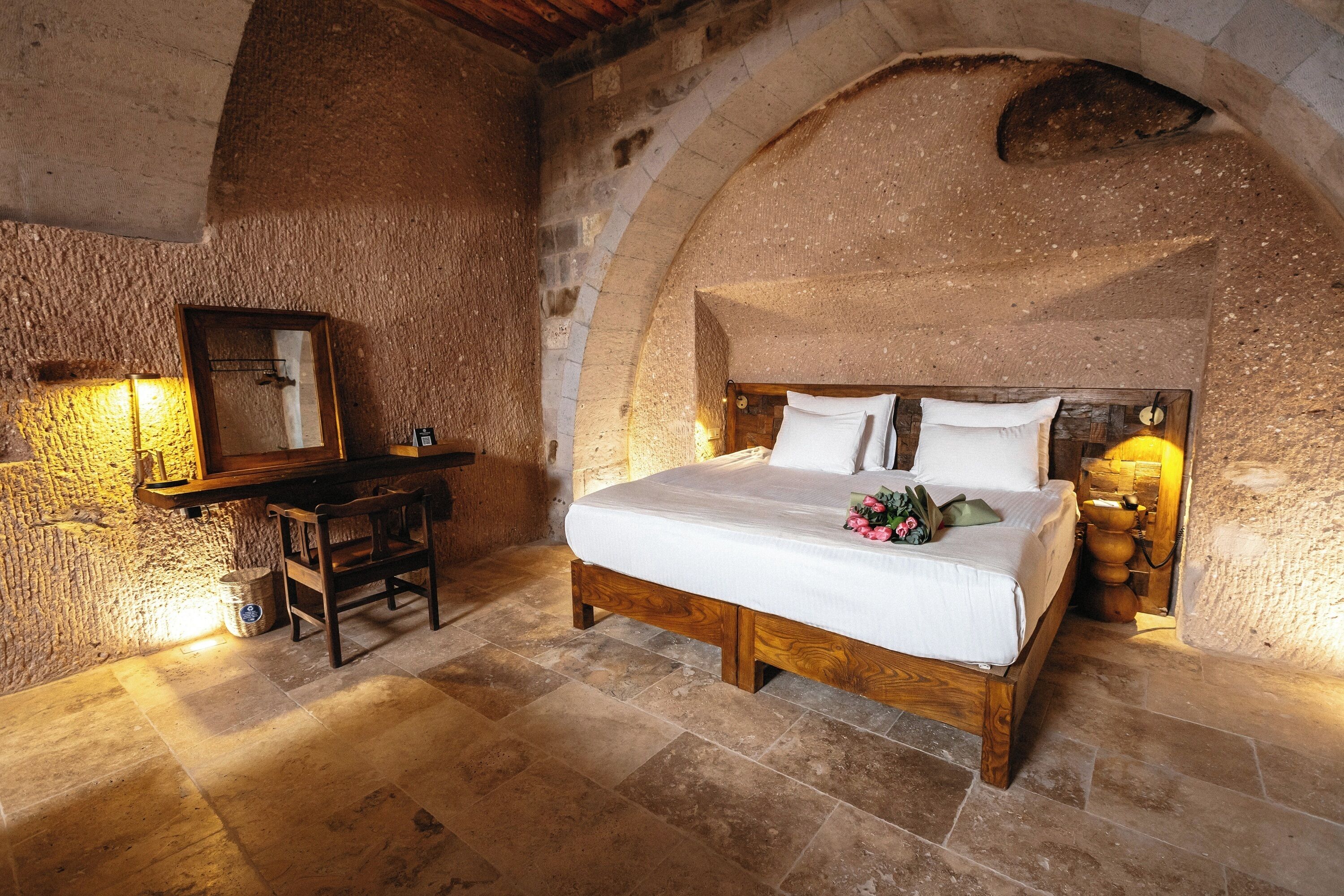 A bedroom with a stone wall, a wooden bed, a desk, and a mirror.
