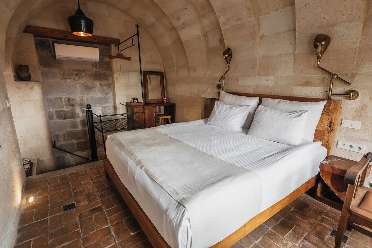 A bedroom with a large bed, stone walls, a wooden bench, and a hanging light fixture.