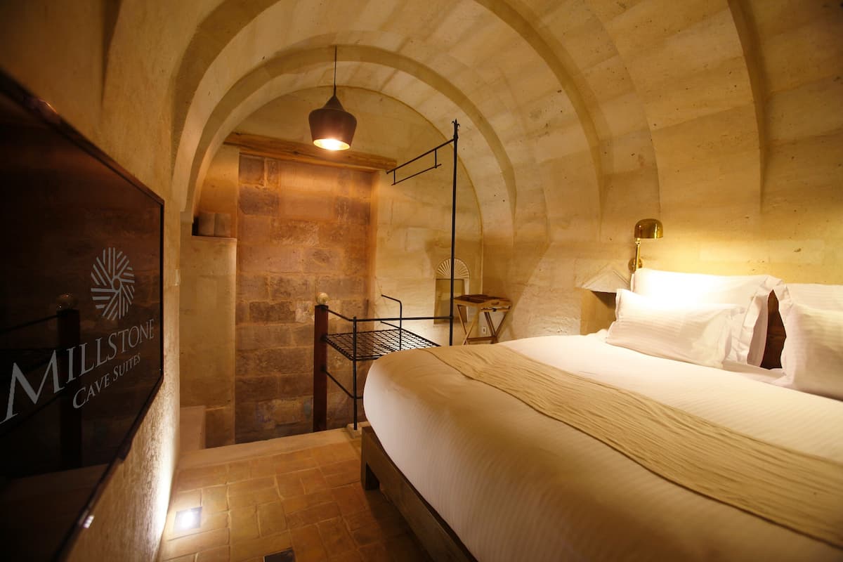 A hotel room with a bed, a nightstand, a wall-mounted lamp, an arched ceiling, and a sign for 'Millstone Cave Suites.'