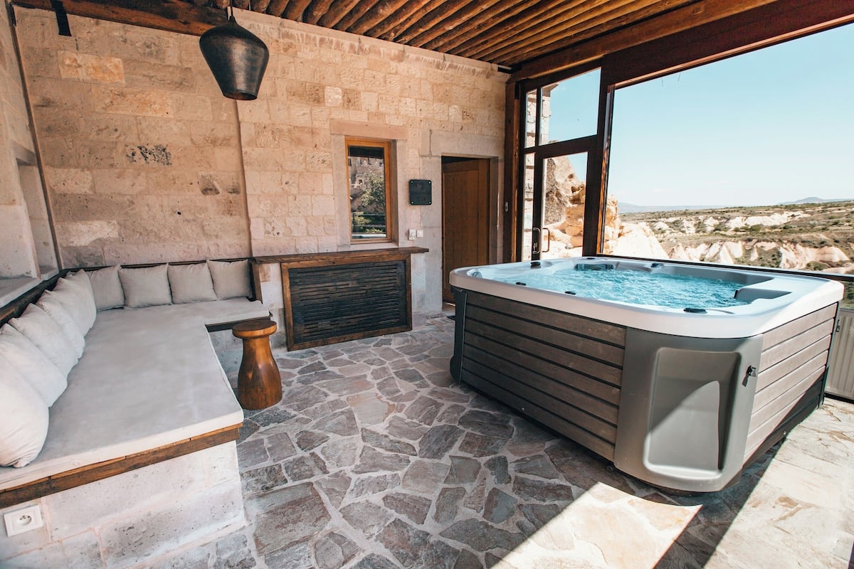 A stone-floored spa area with a hot tub, a cushioned seating area, and a view of the surrounding landscape.