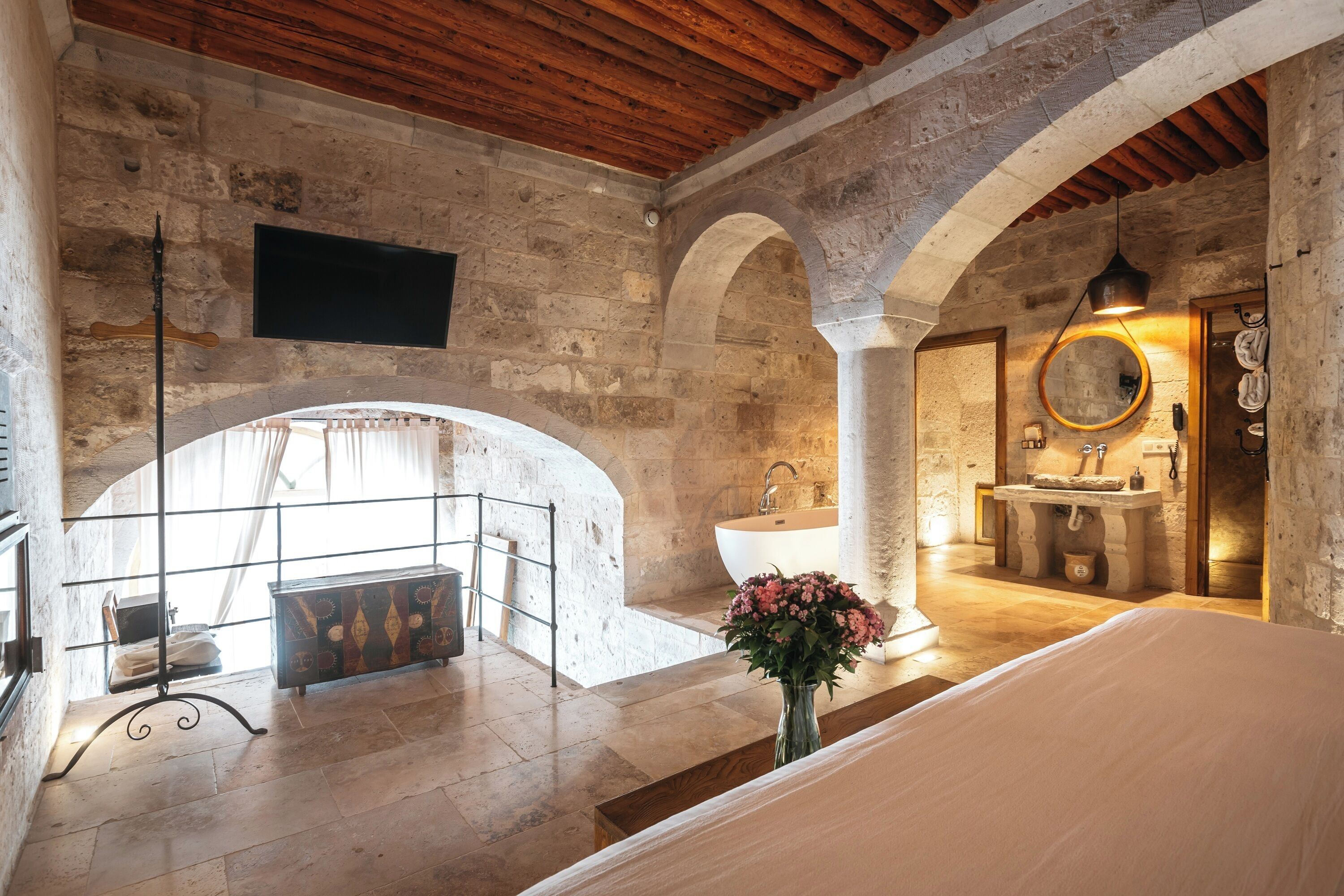 Ein Schlafzimmer mit einem großen Bett, Holzboden, Steinwänden, einem Fernseher, einem Badezimmer mit Waschbecken und Spiegel sowie einem Balkon mit Geländer.