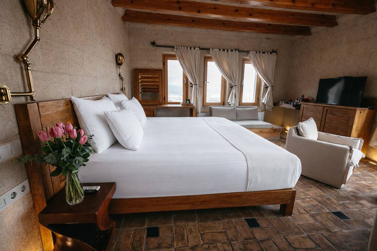A bedroom with a large bed, a wooden headboard, a bedside table with a vase of flowers, a wooden bench, a sofa, and a television.