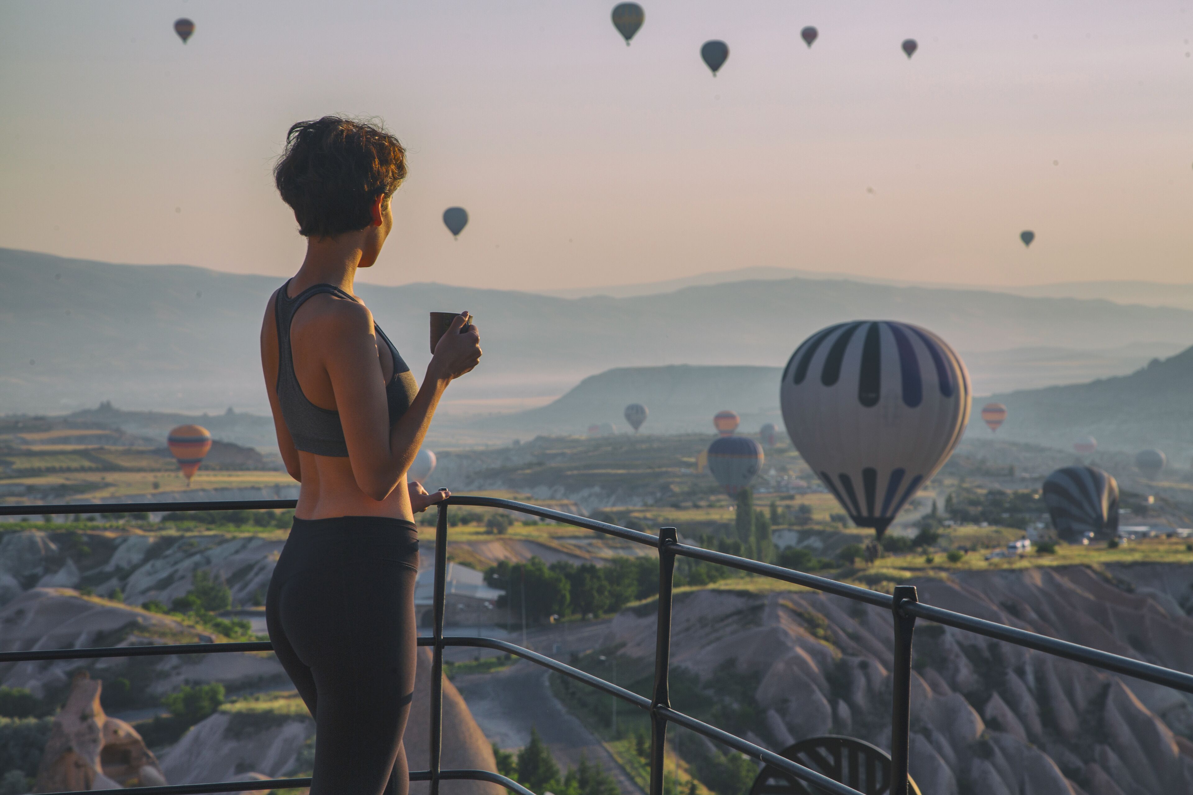 A person standing by a railing overlooking a landscape with multiple hot air balloons.