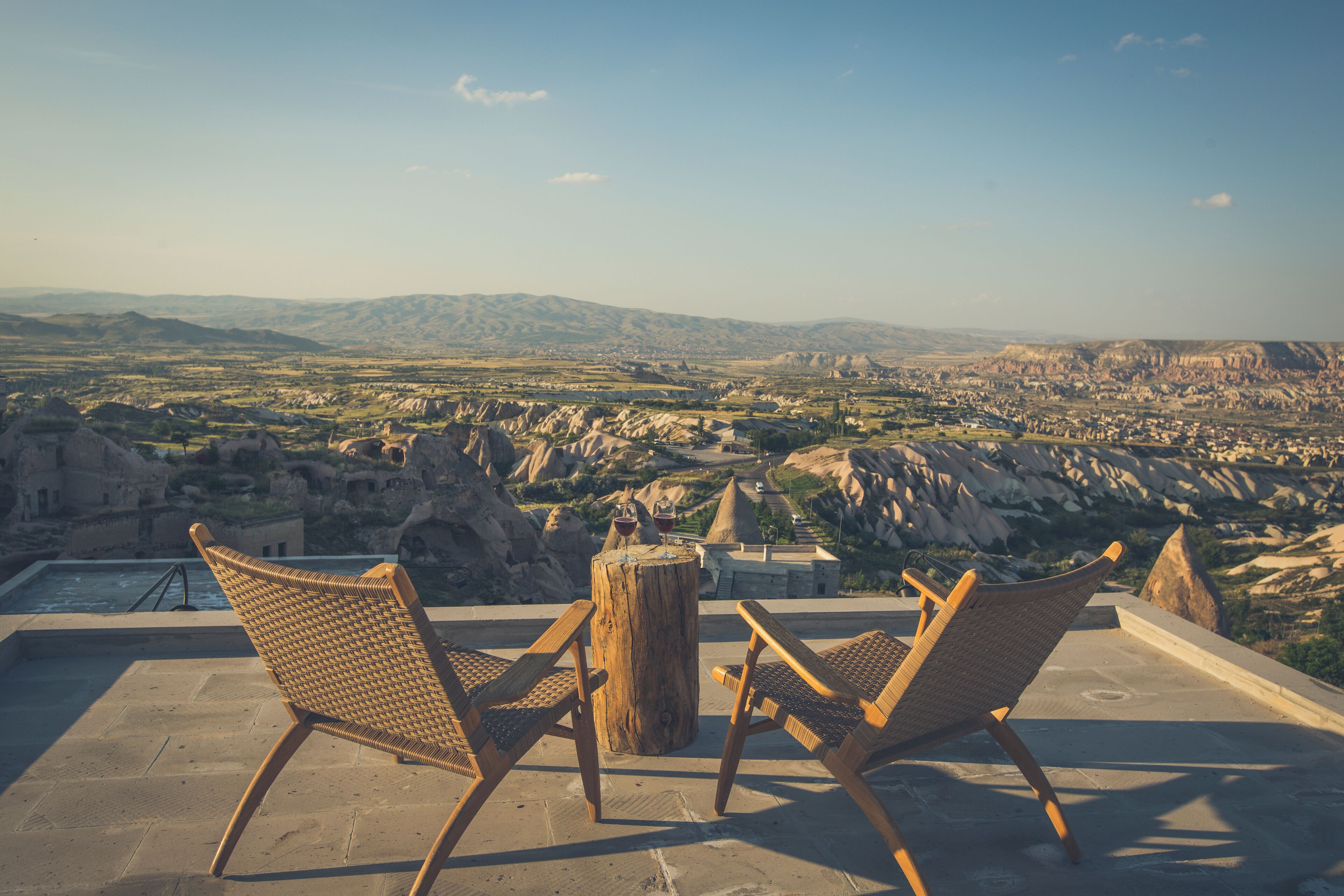 A terrace with wicker chairs and a wooden table, overlooking a mountainous landscape with a village below.