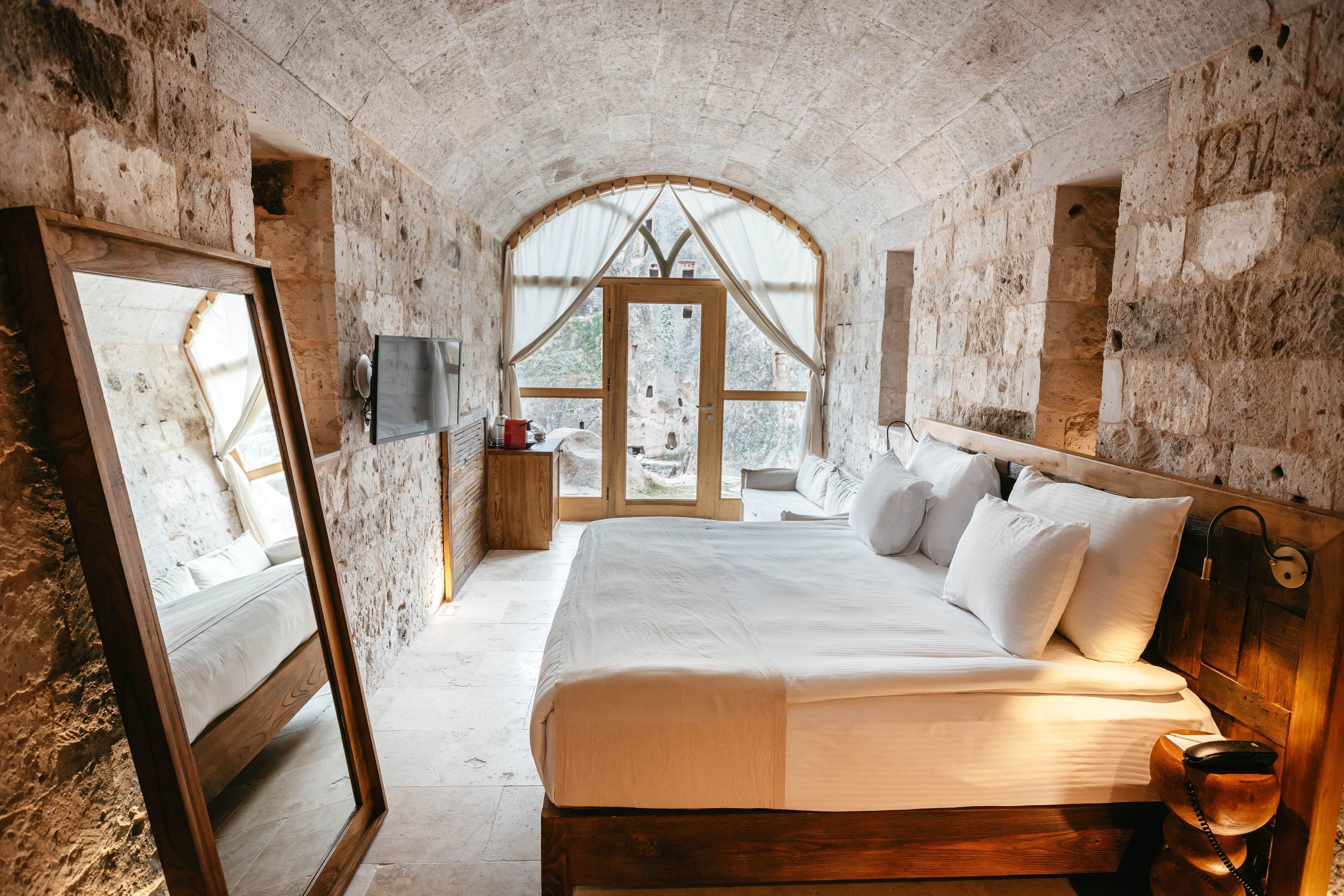 A bedroom with a large bed, wooden headboard, stone walls, a wooden door, and a TV on the wall.