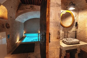 A stone-walled bathroom with a bathtub, a wooden bench, a stone sink, and a round mirror with a light above it.