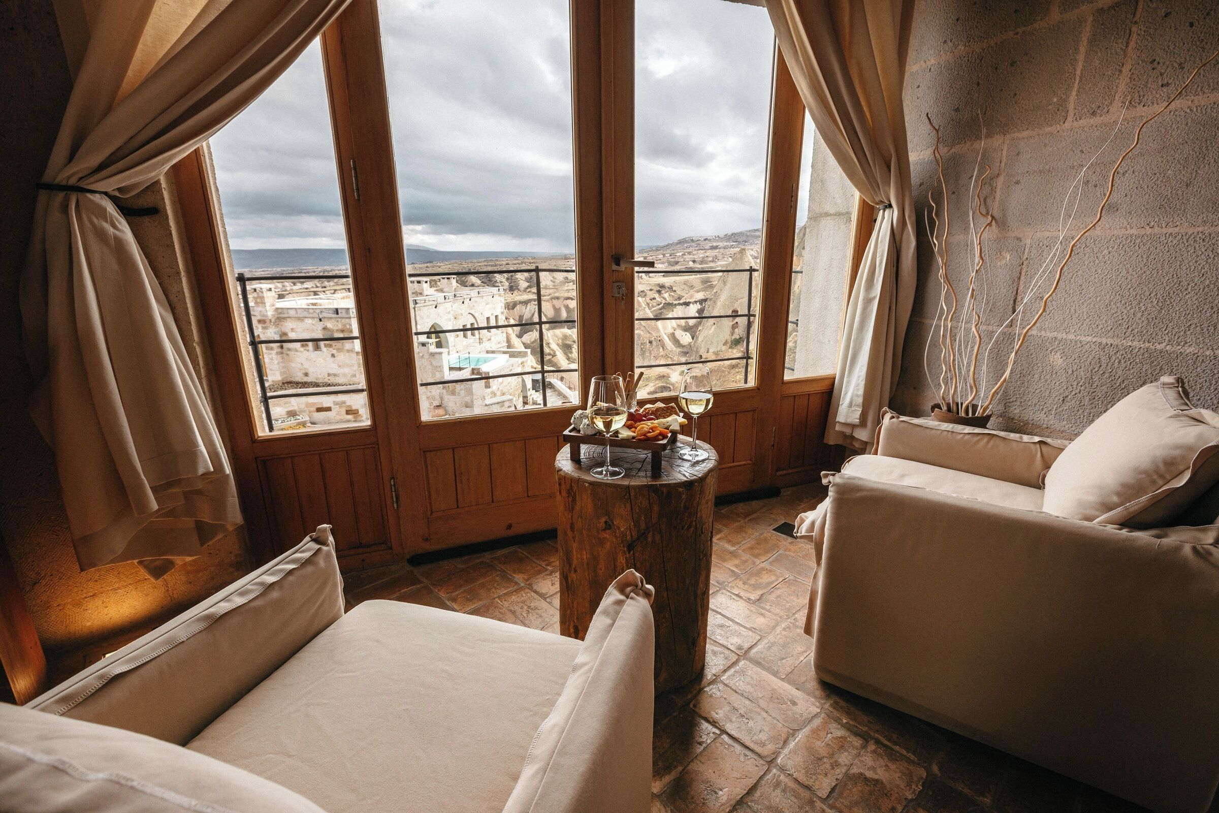 Quarto com uma cama, duas cadeiras, uma mesa de madeira com um copo de vinho e vista para uma paisagem distante através de grandes janelas.